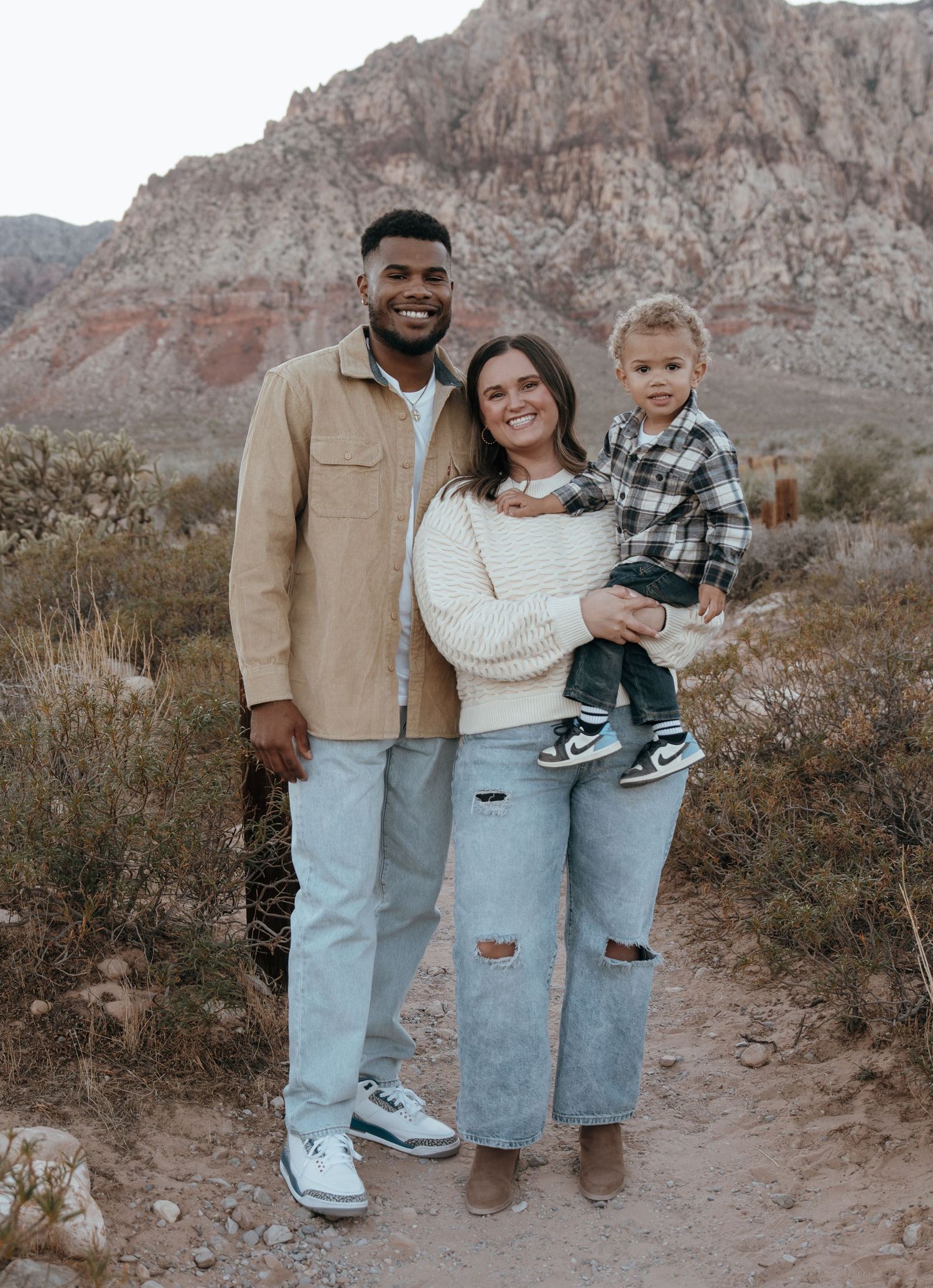 A happy family of three poses on a desert trail with red rock mountains in the background.
