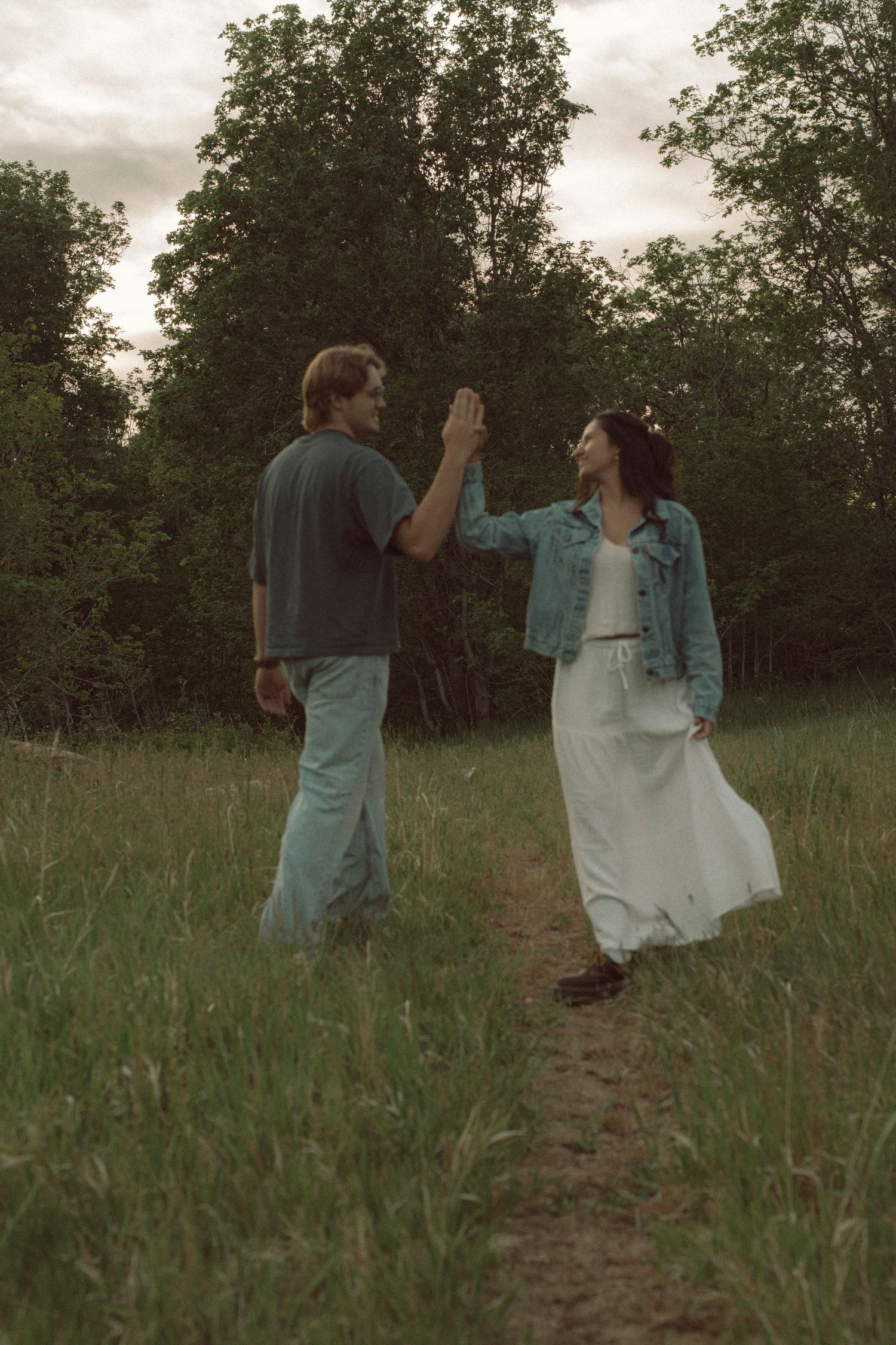 Couple high-fiving on a grassy path surrounded by lush green trees, woman in white dress and denim jacket.
