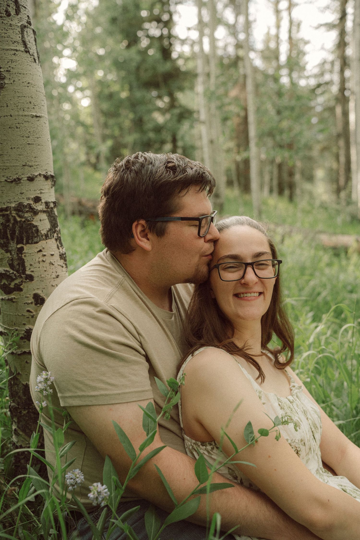 A couple sits together in a forest meadow, the man kissing the woman's cheek as she smiles warmly.