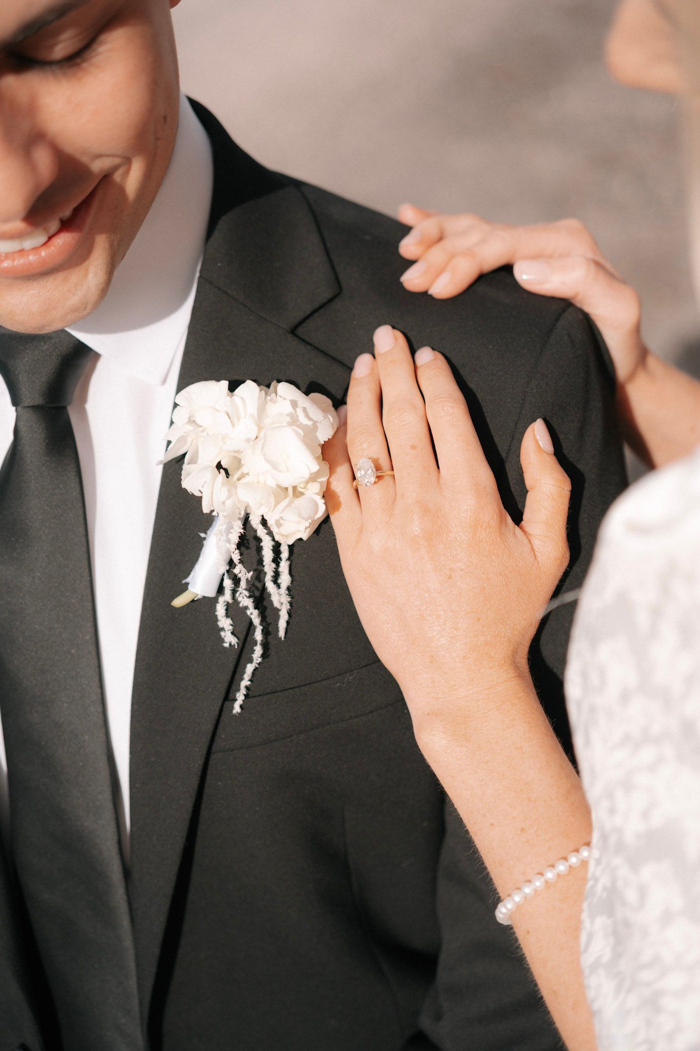 Bride places hand on grooms black suit lapel showing diamond engagement ring and pearl bracelet near boutonniere.