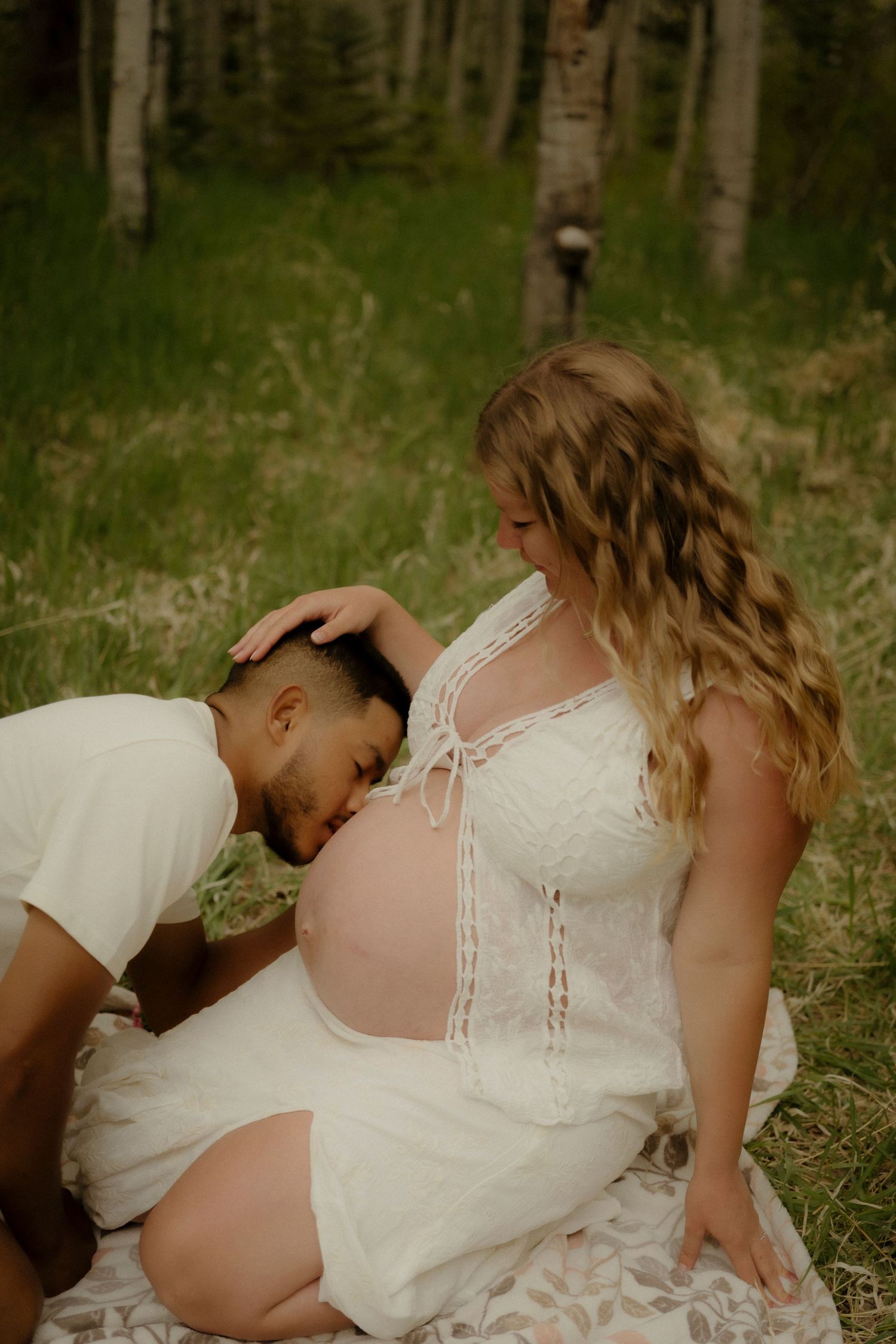 A man tenderly kisses a pregnant woman's bare belly during an outdoor maternity photo session in a forest.
