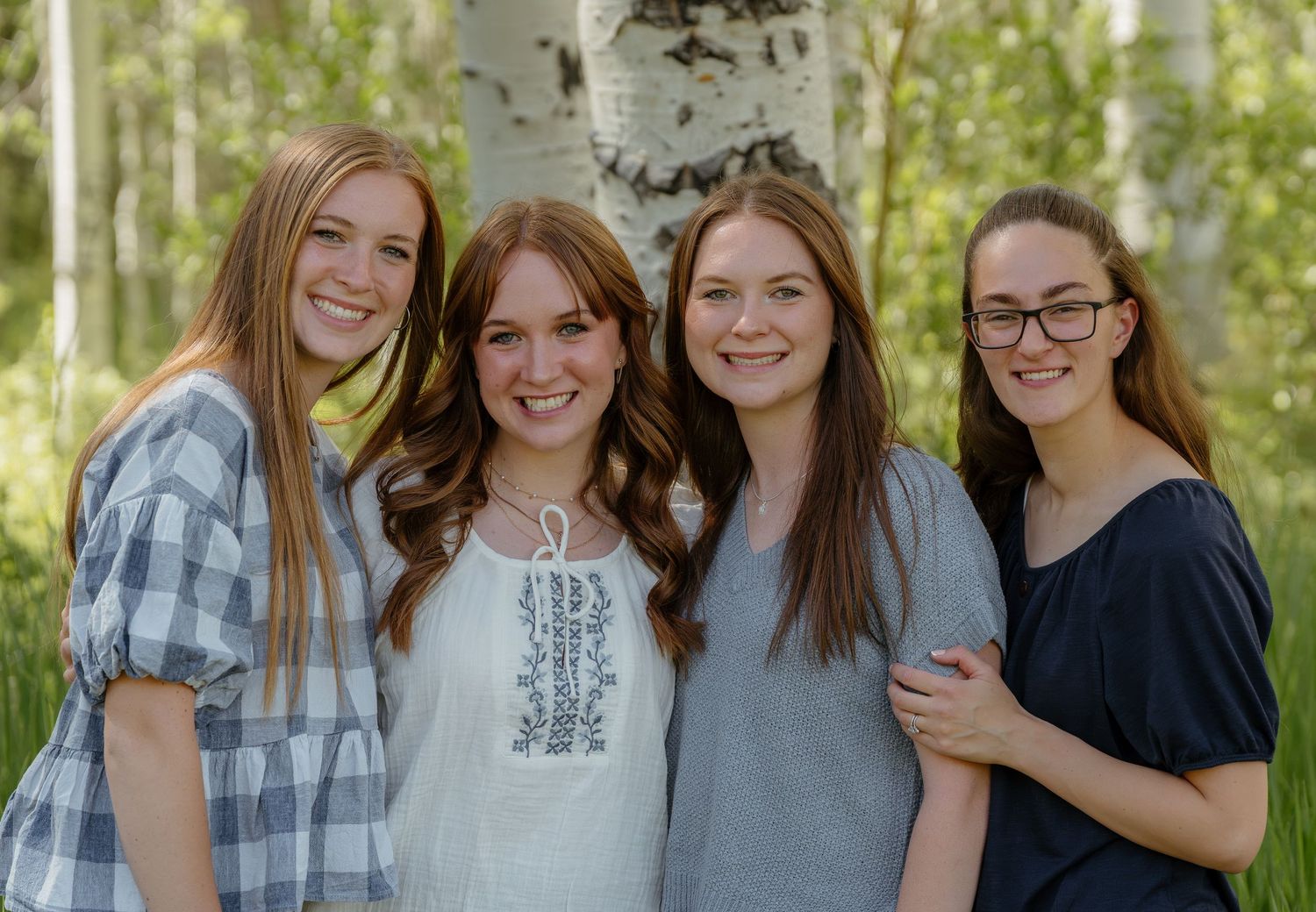 Four smiling young women pose together outdoors among white birch trees in a bright, cheerful family portrait.