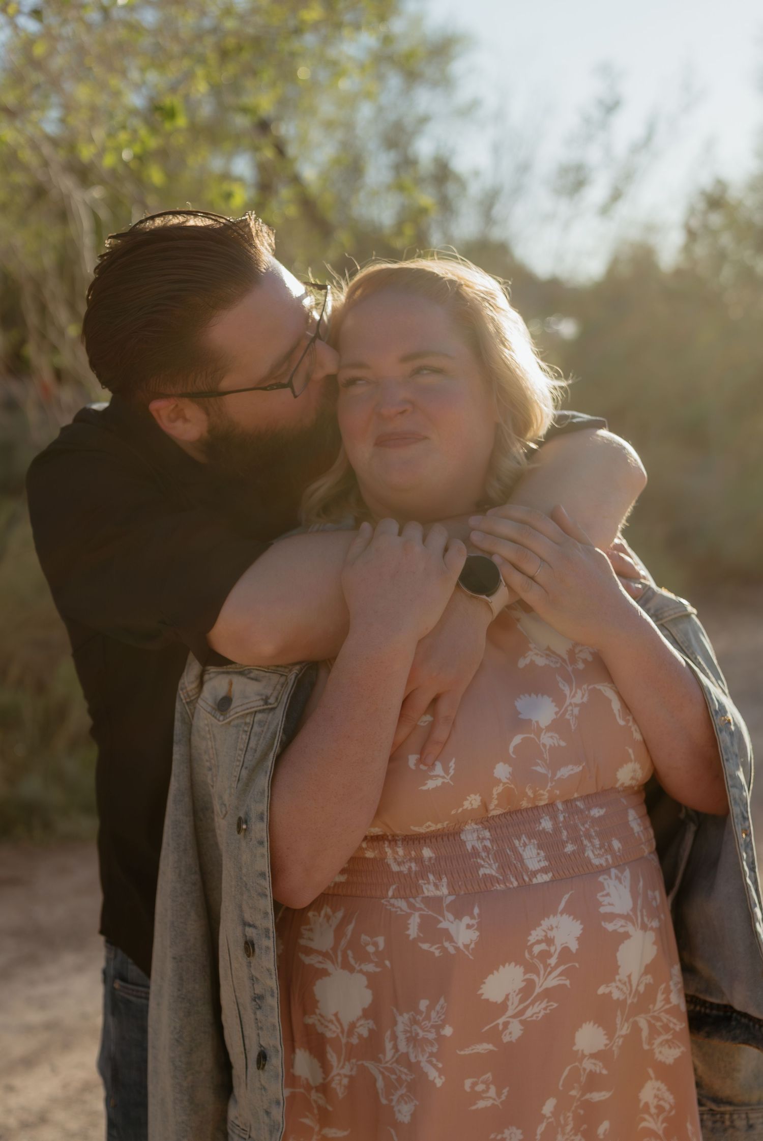 A couple shares a tender moment outdoors, man kissing woman's cheek while embracing her in warm golden sunlight.