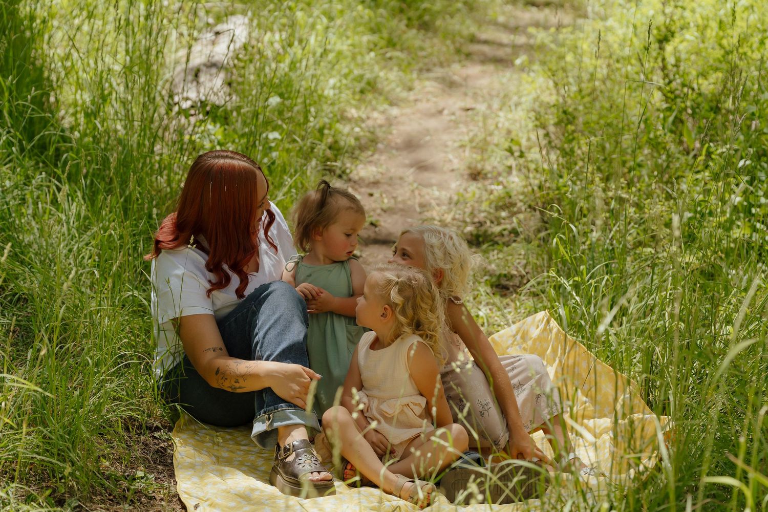 A woman with red hair sits on a blanket in tall grass with two young children during an outdoor picnic.