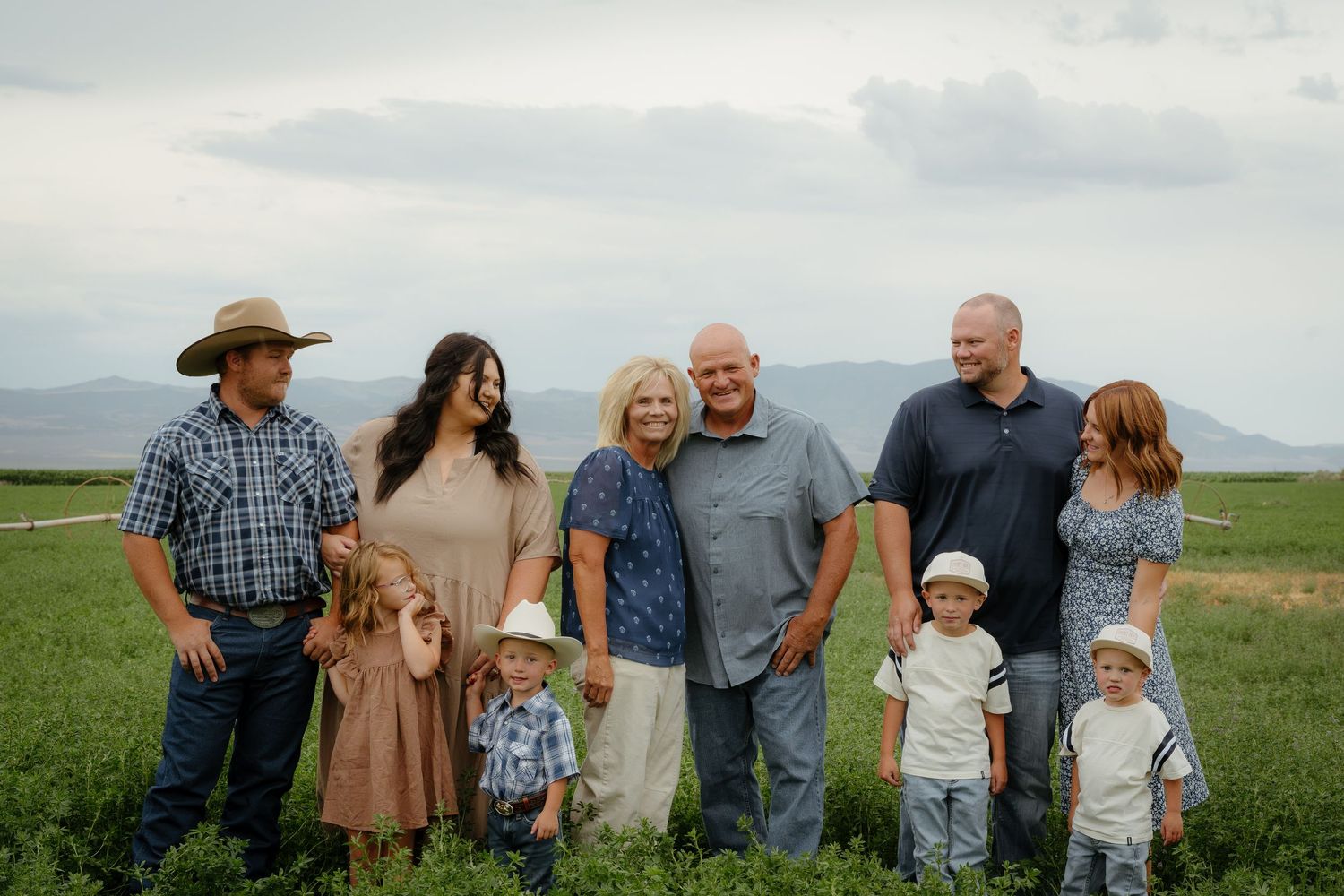 Multi-generational family portrait in green farm field with mountain backdrop on cloudy day.