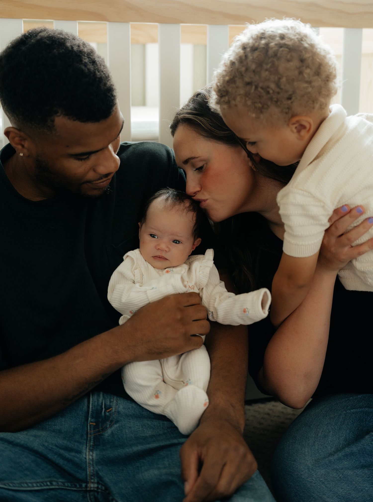 A loving interracial family of four cuddles together, with parents kissing their toddler while holding their newborn baby.