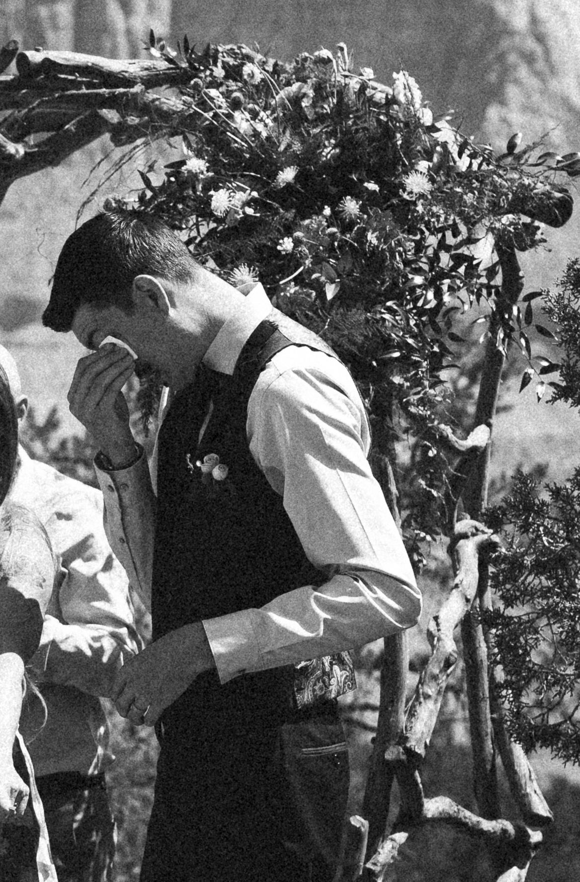 Emotional groom in vest wiping tears under floral arch at outdoor wedding ceremony, black and white photo.