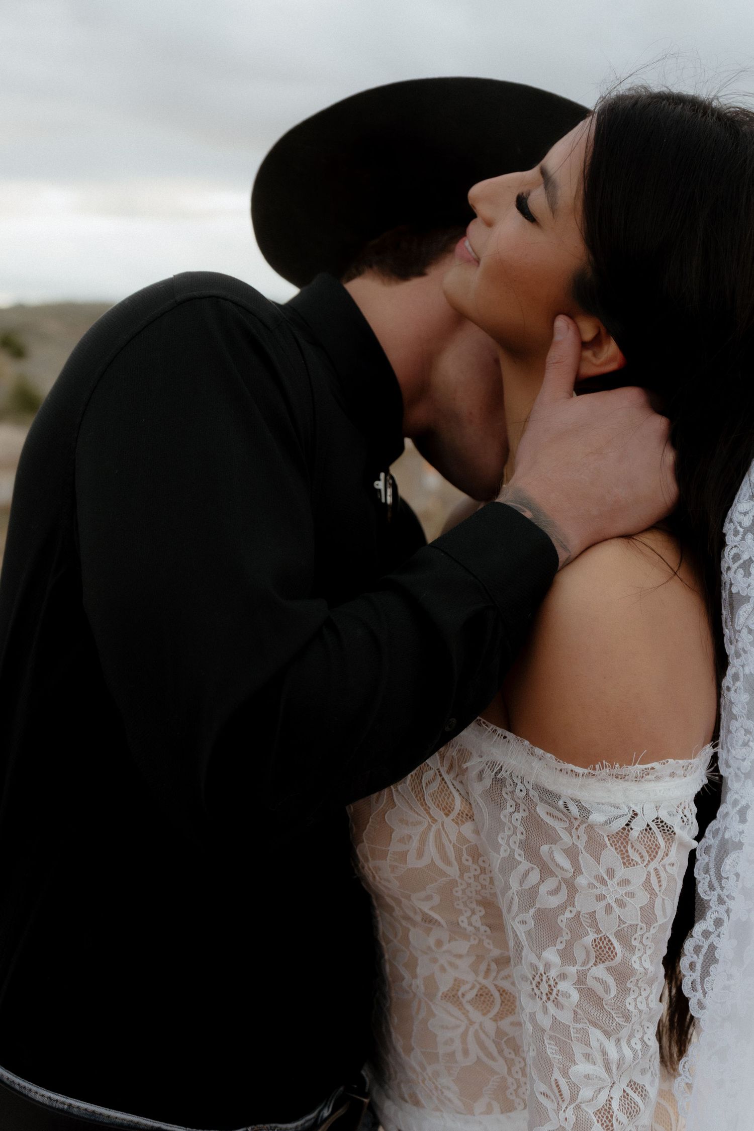 A bride in white lace dress and groom in black cowboy hat share a romantic kiss outdoors.