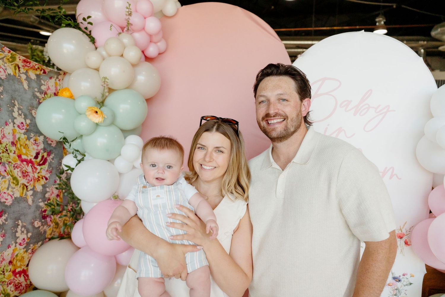 A couple holding a baby at a pastel pink and green balloon-decorated baby shower backdrop.