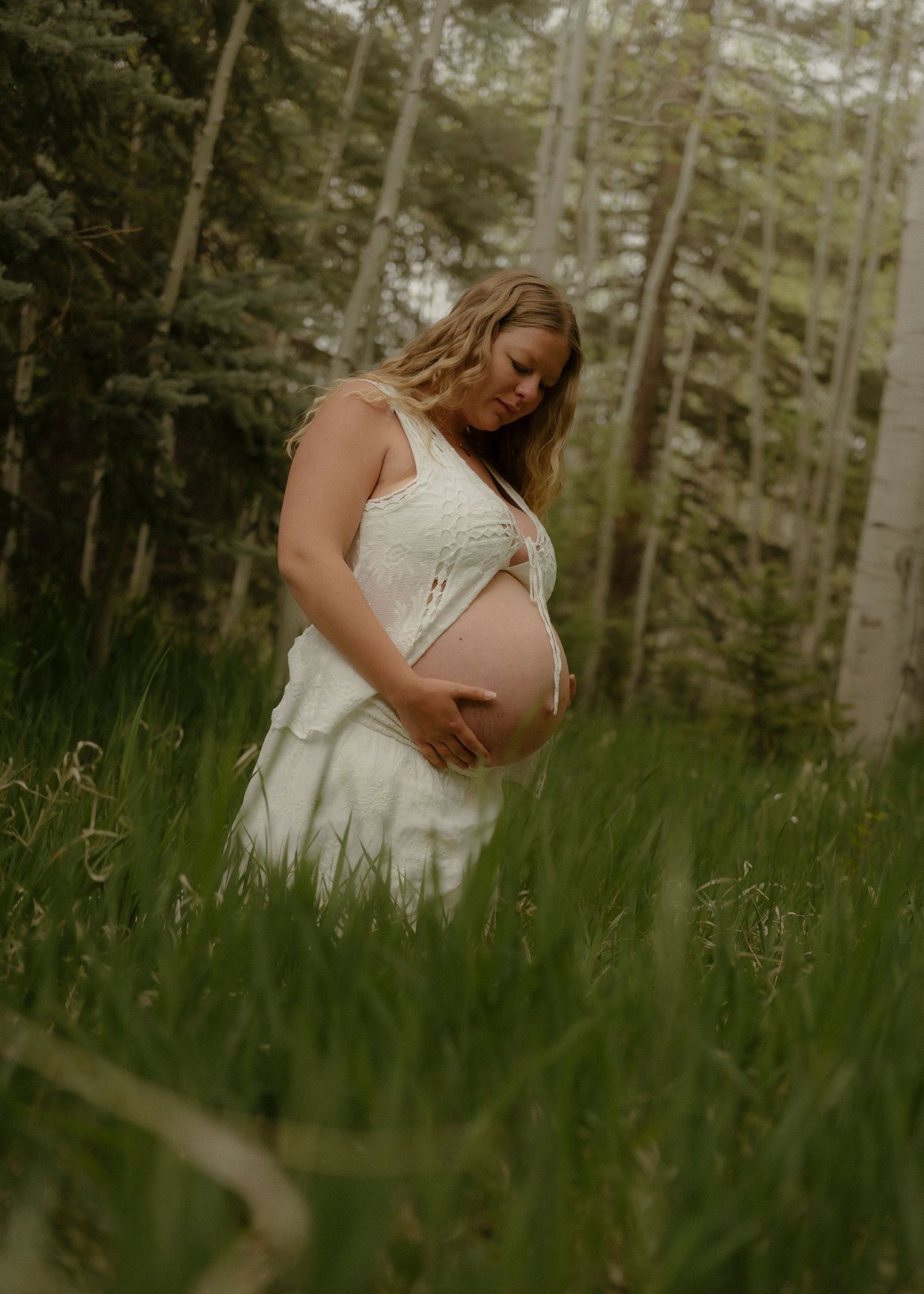Pregnant woman in white dress cradling baby bump in lush green meadow surrounded by tall trees.