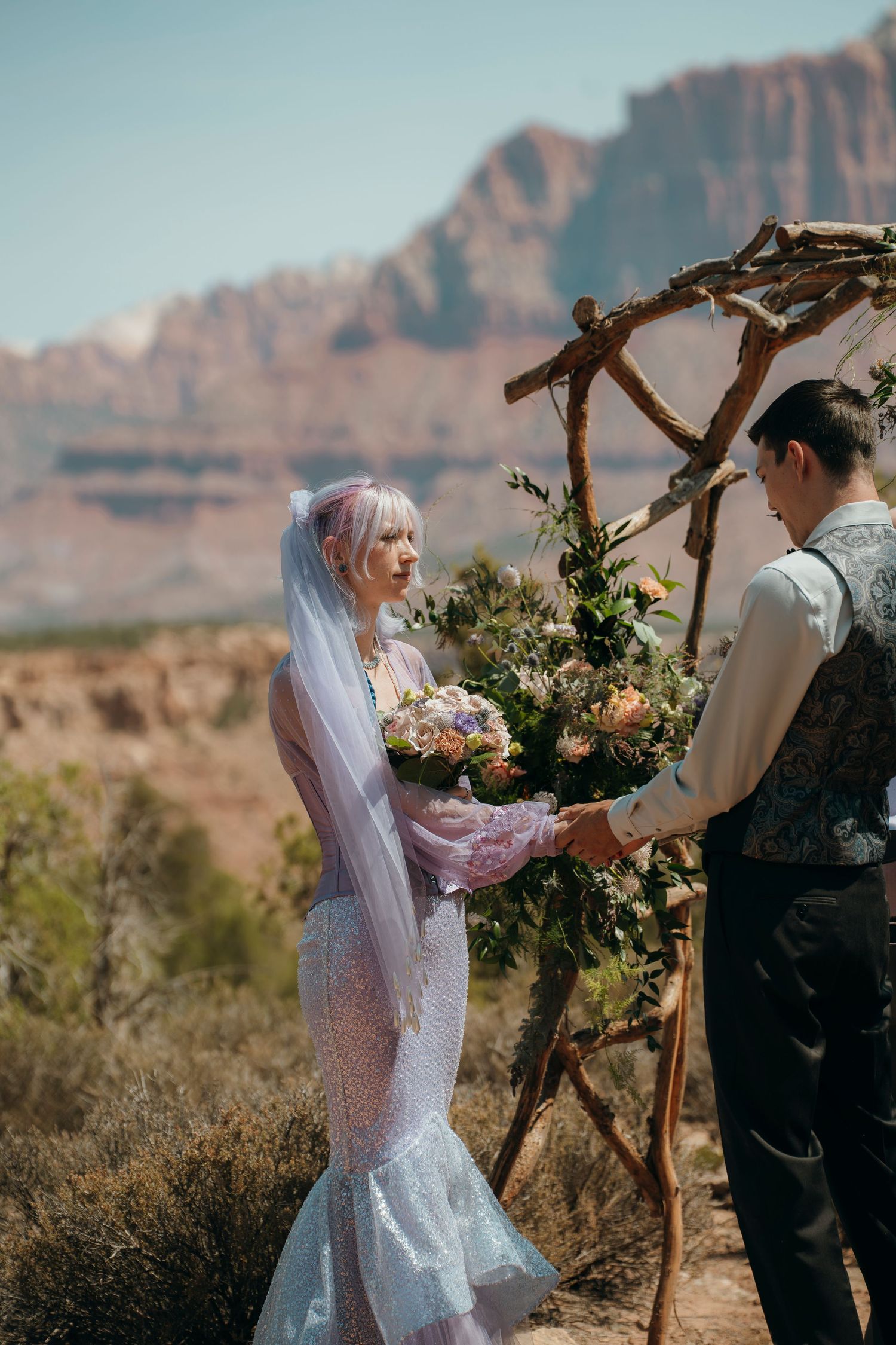 Bride with pink hair and veil holds bouquet with groom at rustic arch, red rock canyon backdrop.