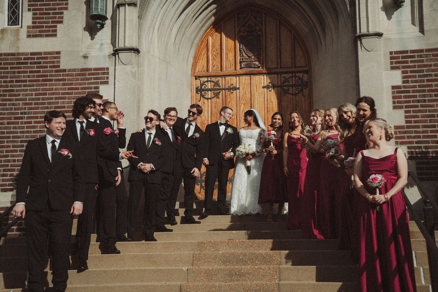 Wedding party poses on church steps, groomsmen in black suits and bridesmaids in burgundy gowns flanking the bride and groom.