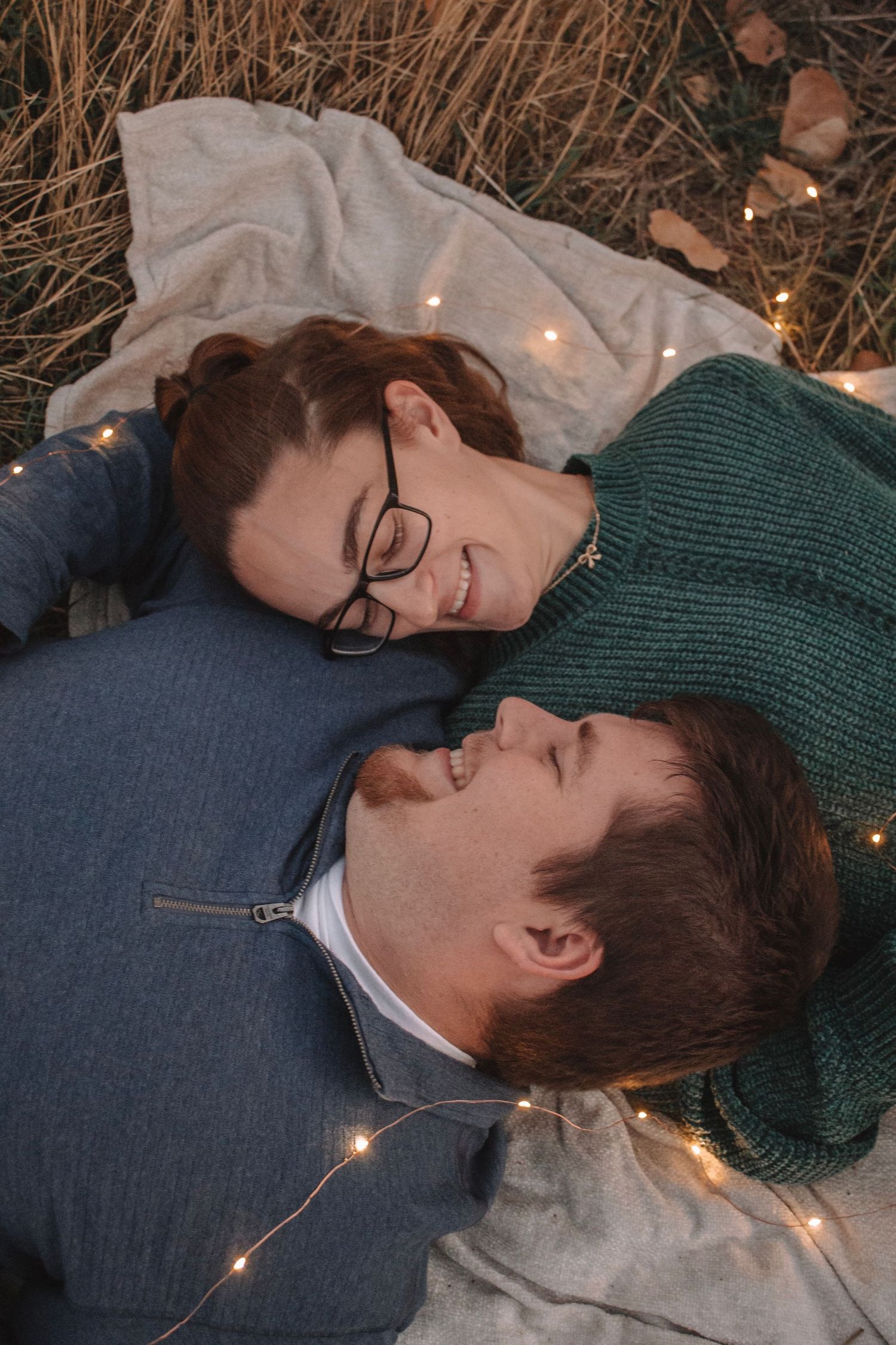 Couple lying on blanket surrounded by fairy lights in dry grass, smiling at each other romantically.
