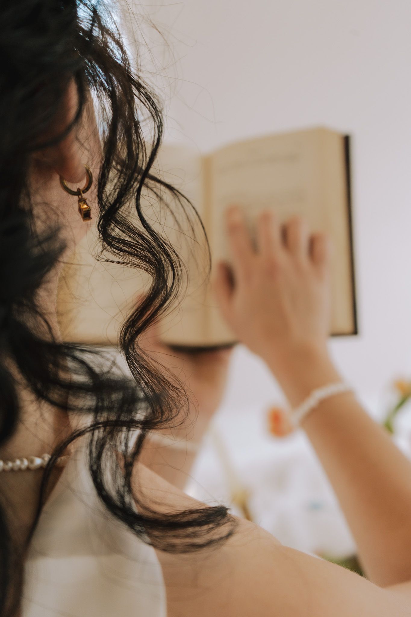 Bride with dark curly hair wearing pearl necklace and gold earring reads a book on her wedding day.