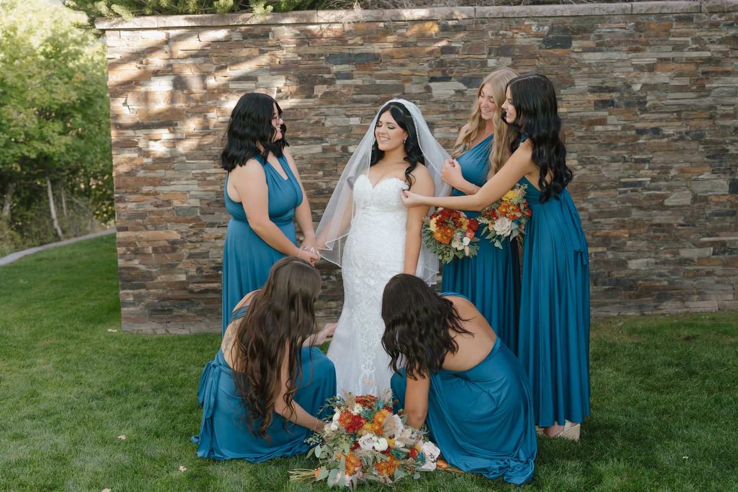Bride in white lace gown surrounded by bridesmaids in teal dresses holding orange bouquets against stone wall backdrop.