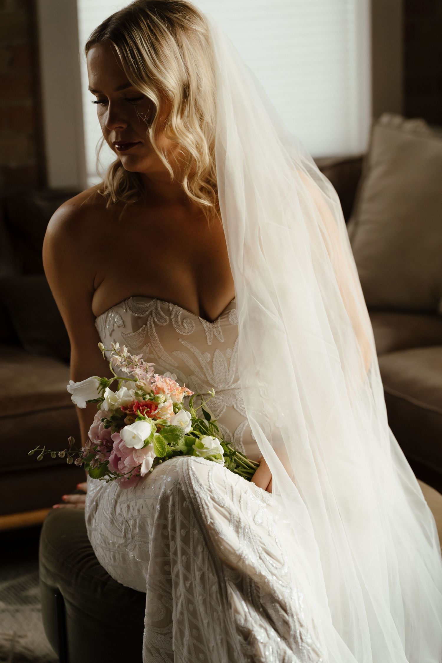 Bride in strapless lace gown and veil holding pink and white bouquet, seated in soft natural light.