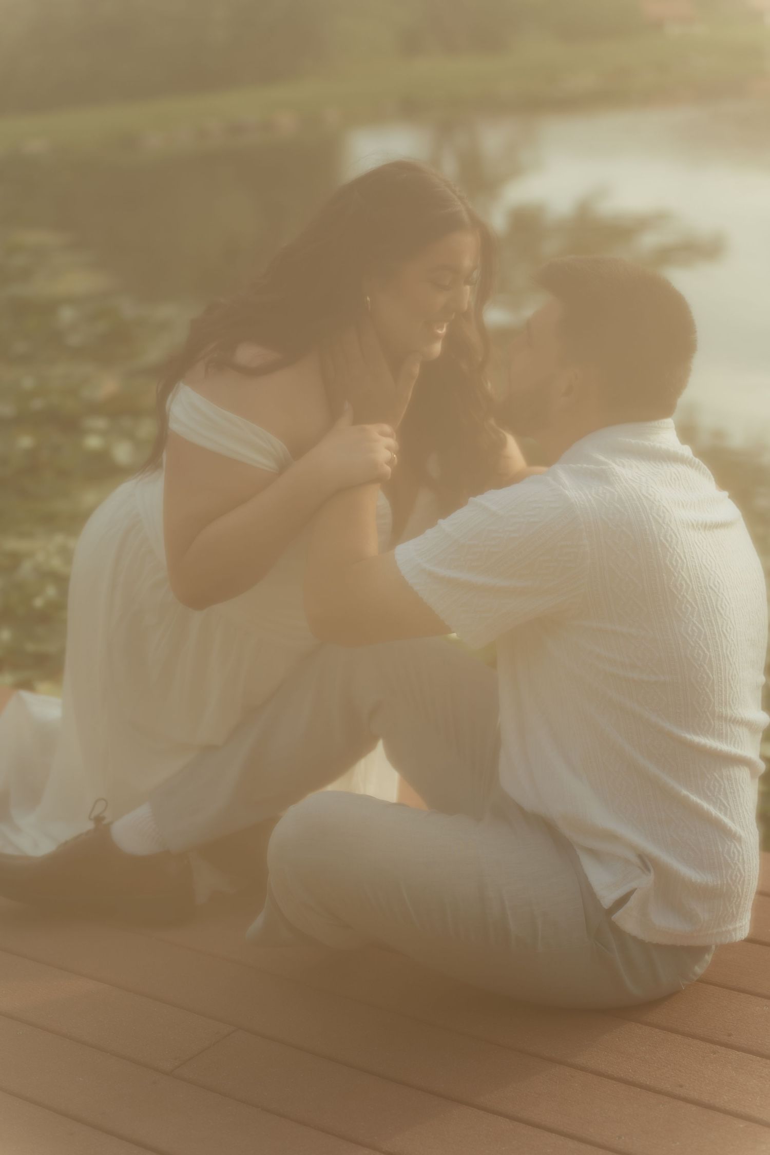 A couple sits together on a wooden deck in golden sunset light, dressed in white, sharing an intimate romantic moment.