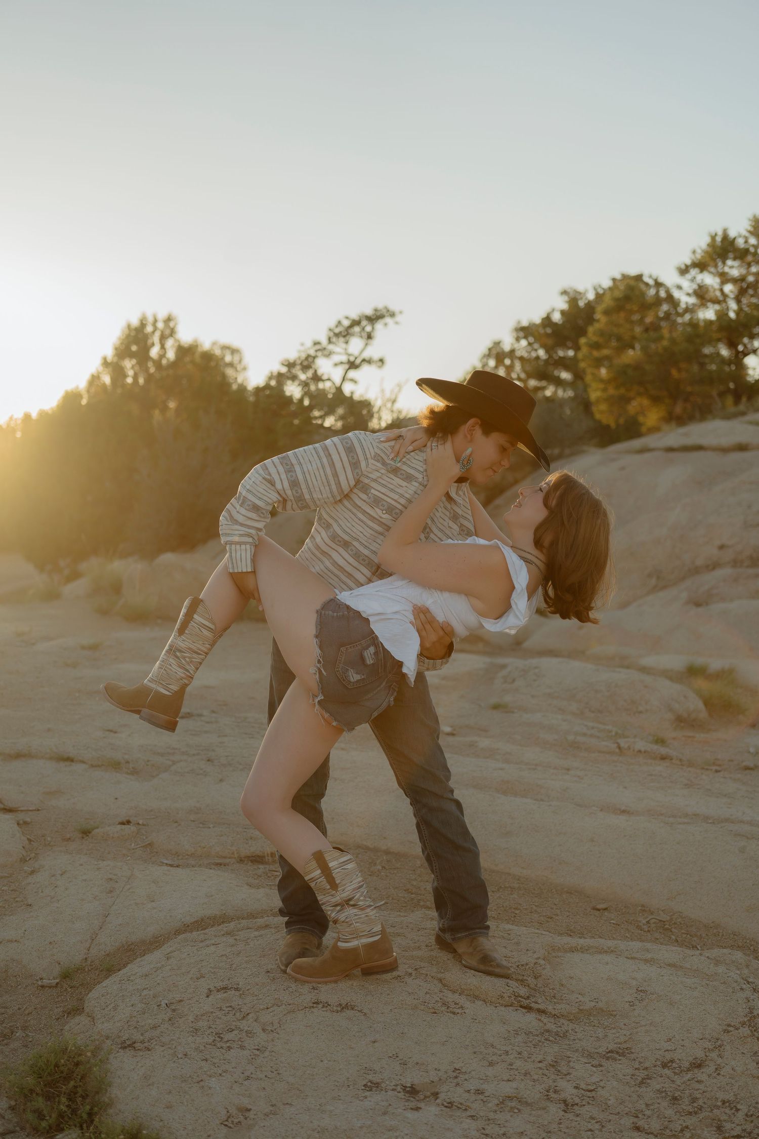 Couple sharing a romantic dip kiss at golden hour in a desert landscape, man in cowboy hat lifting woman.