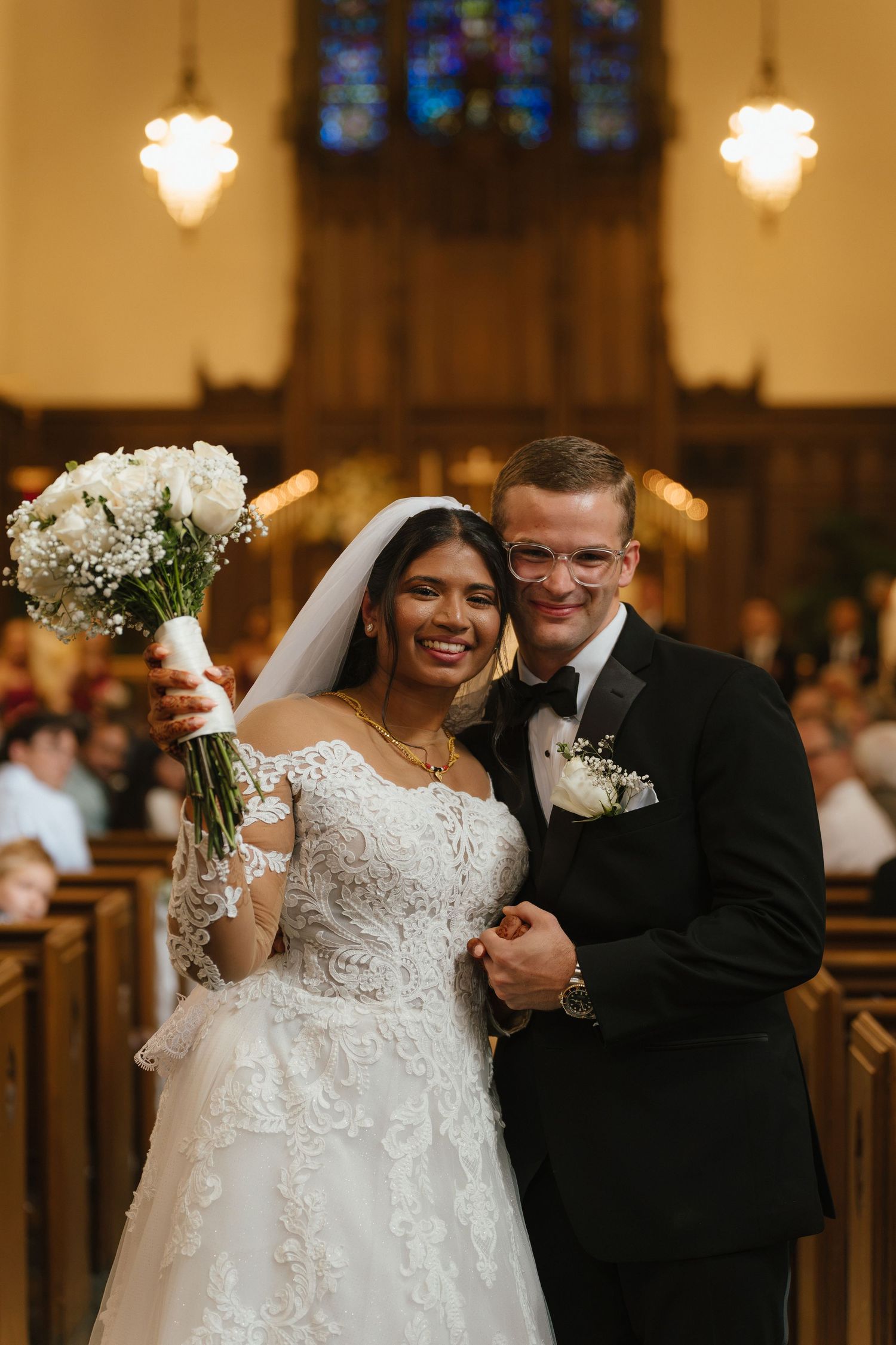 Bride and groom smiling joyfully at church altar, bride holding white rose bouquet in lace gown, groom in black tuxedo.