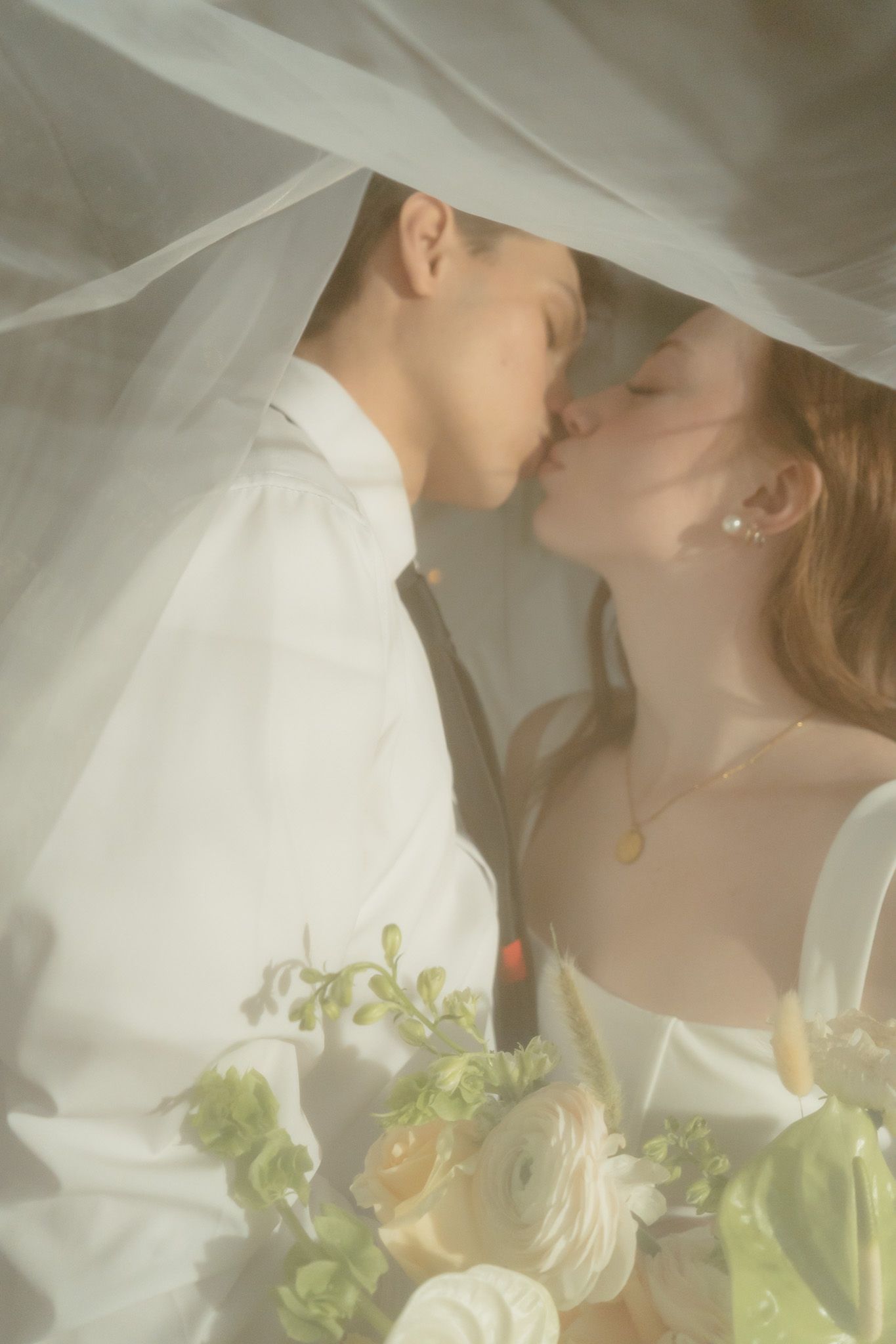 Bride and groom share a tender kiss under a soft white veil with a bouquet of white and green flowers.