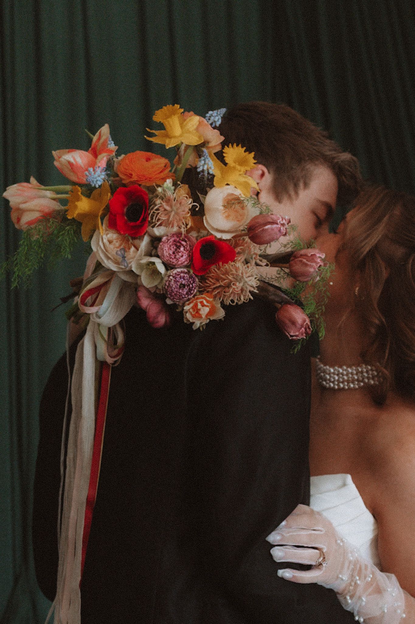 Bride and groom share a romantic kiss hidden behind a vibrant bridal bouquet of red, orange, and pink flowers.