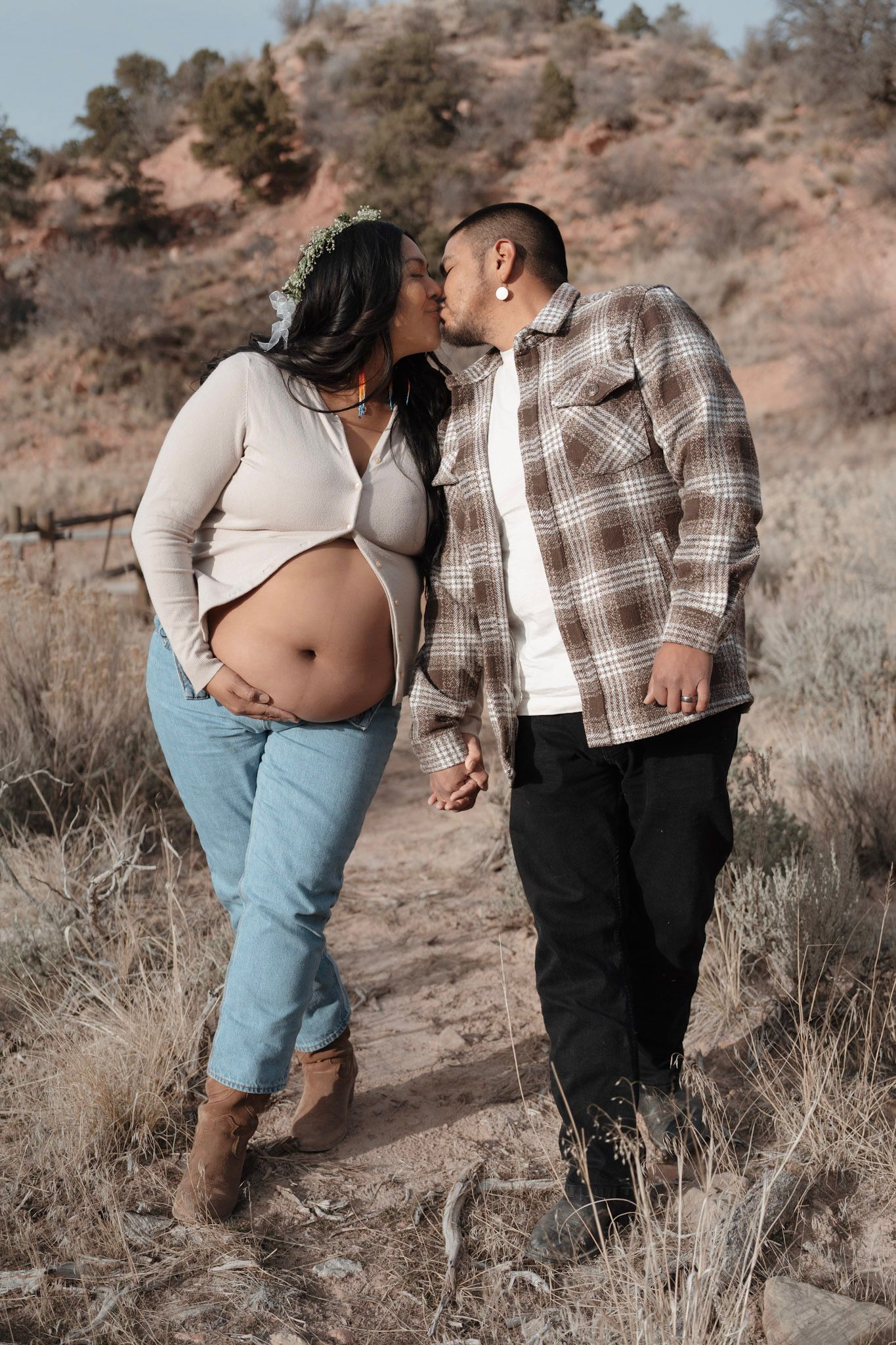 Pregnant couple sharing a kiss outdoors in a desert landscape, woman showing baby bump in jeans and white top.