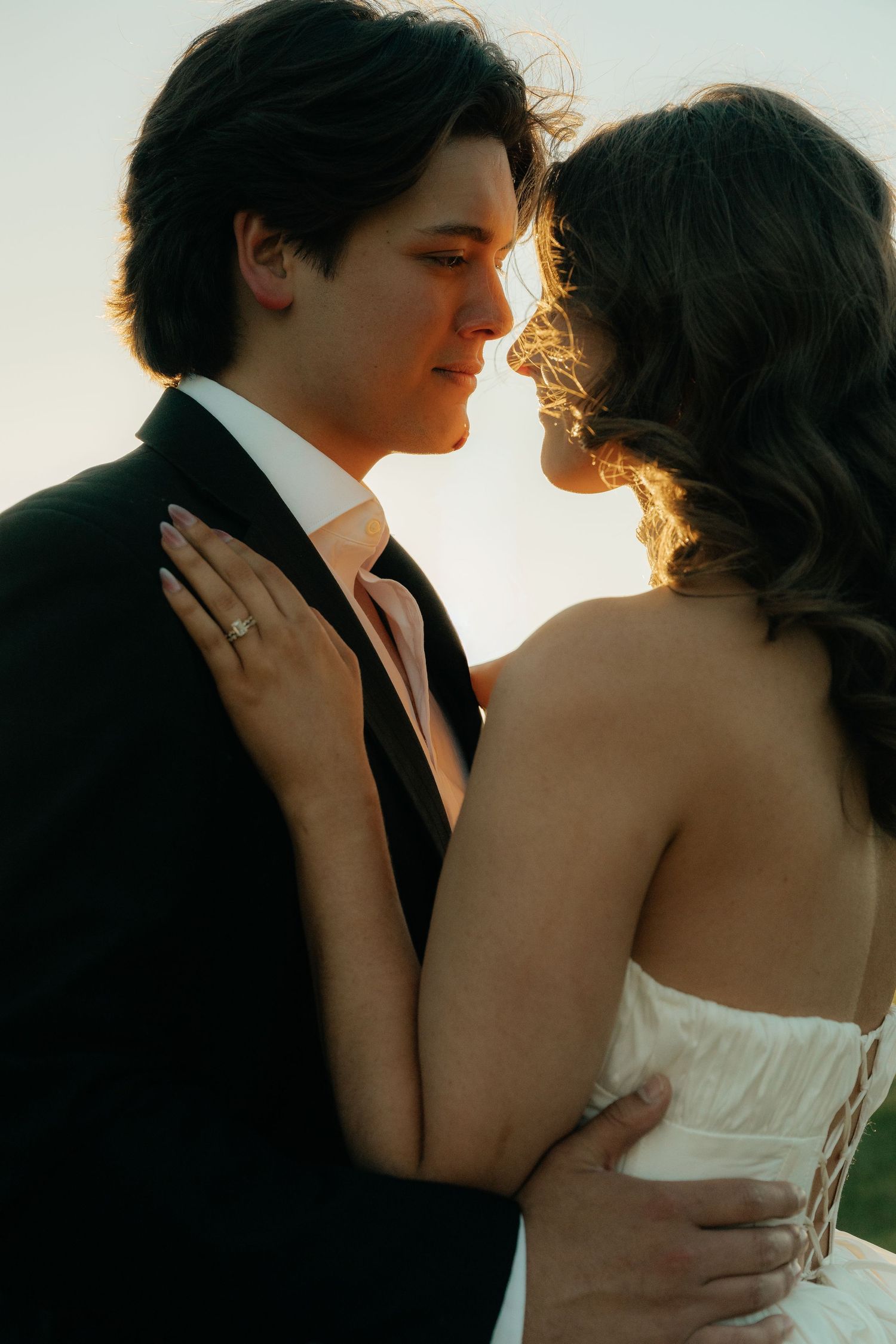 A bride and groom share a romantic moment at sunset, with the bride wearing a white dress and engagement ring.
