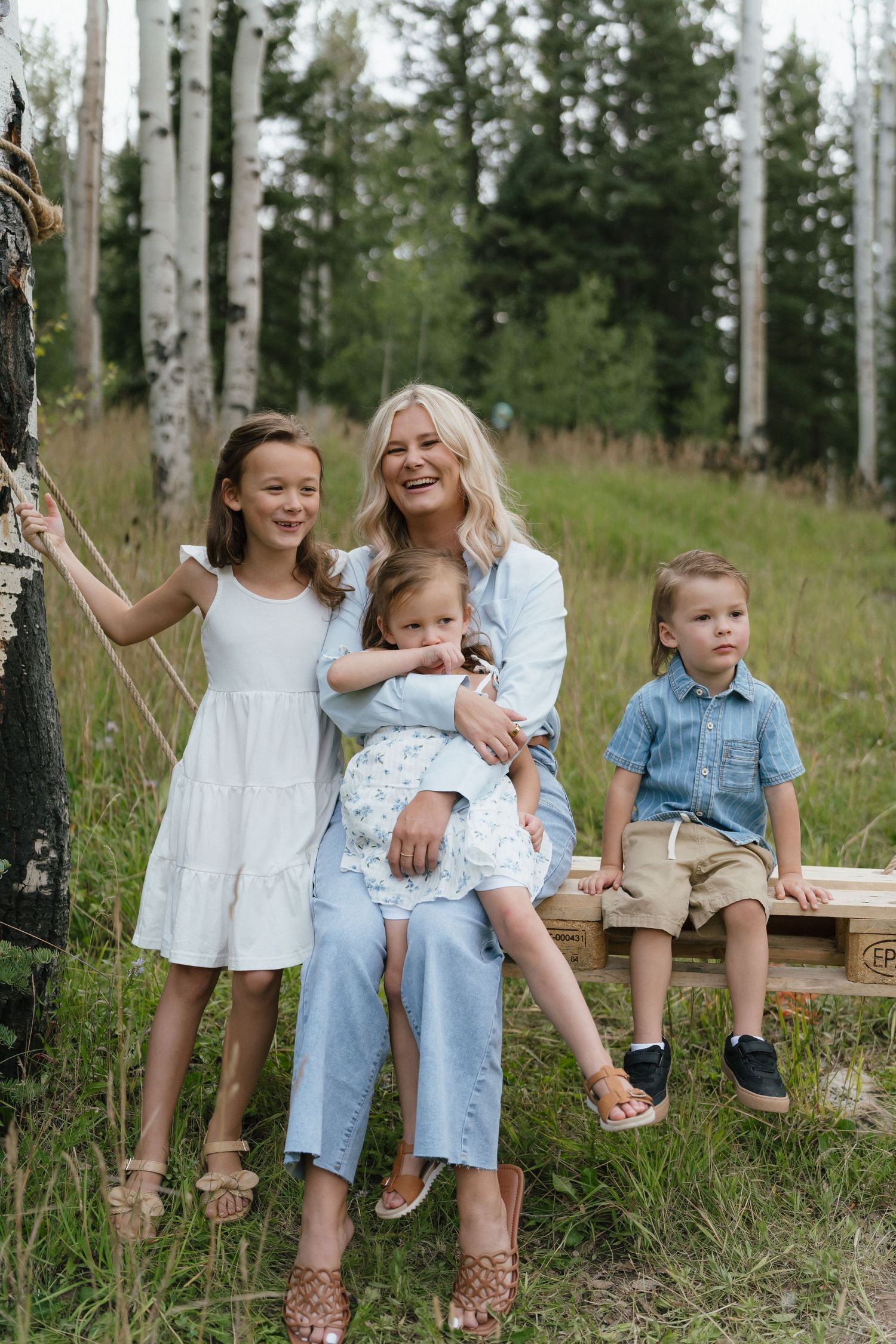 A smiling blonde woman sits outdoors with three young children in a forest clearing, wearing white and blue outfits.