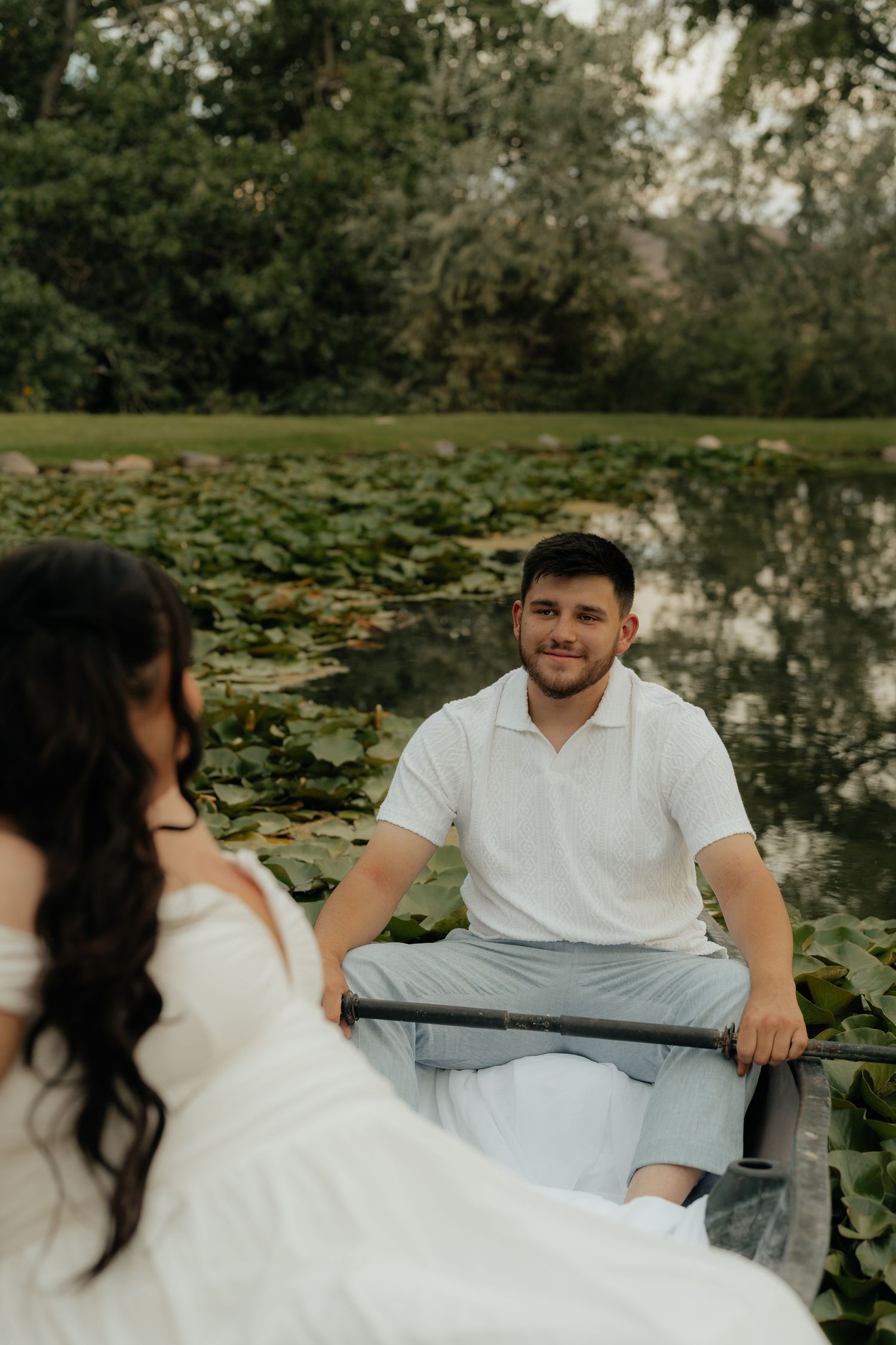 A couple in white attire enjoys a romantic canoe ride on a lily pad-covered pond surrounded by lush greenery.