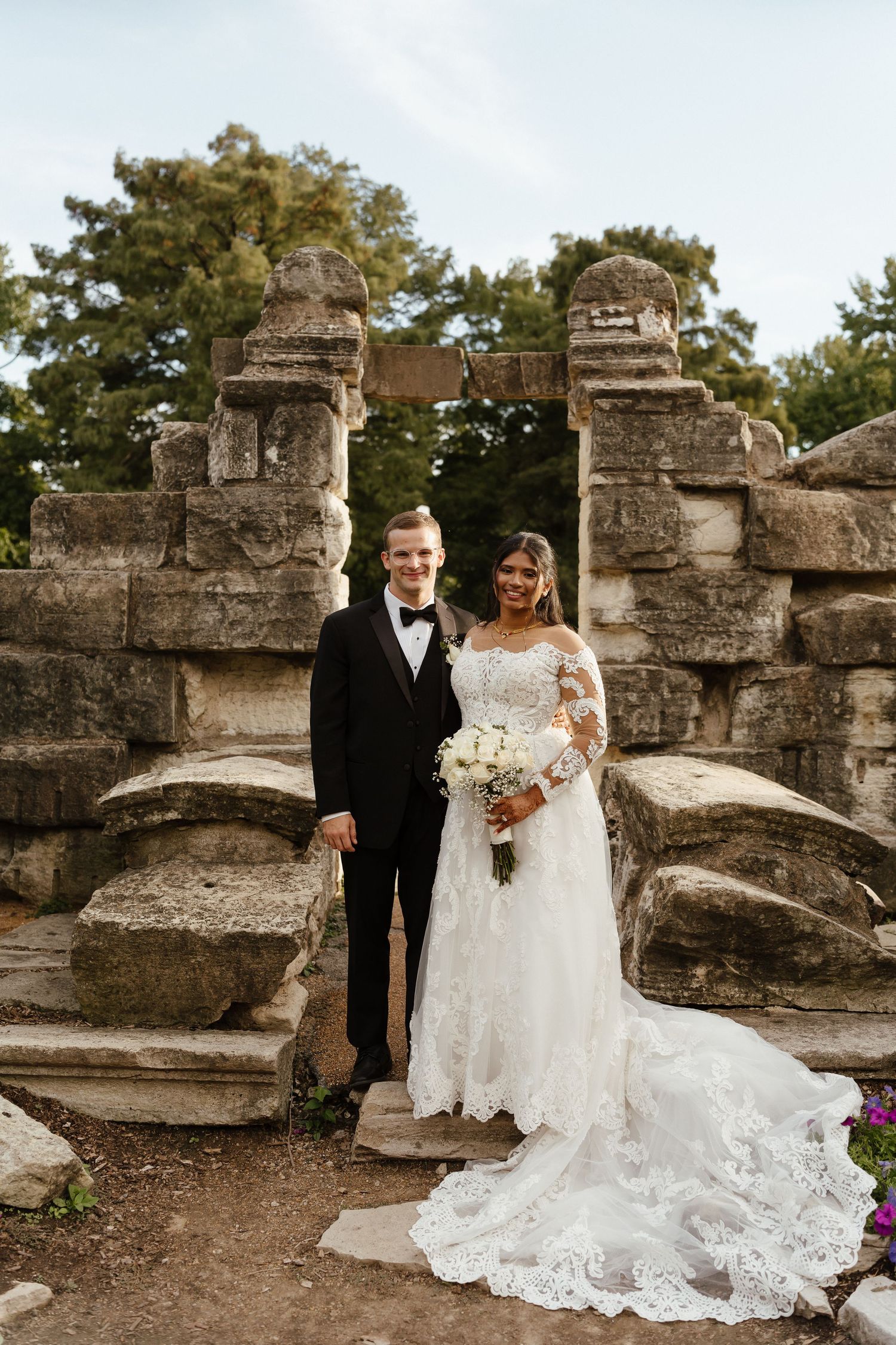 Bride and groom posing in front of ancient stone ruins, bride wearing lace gown holding white bouquet, groom in black tuxedo.