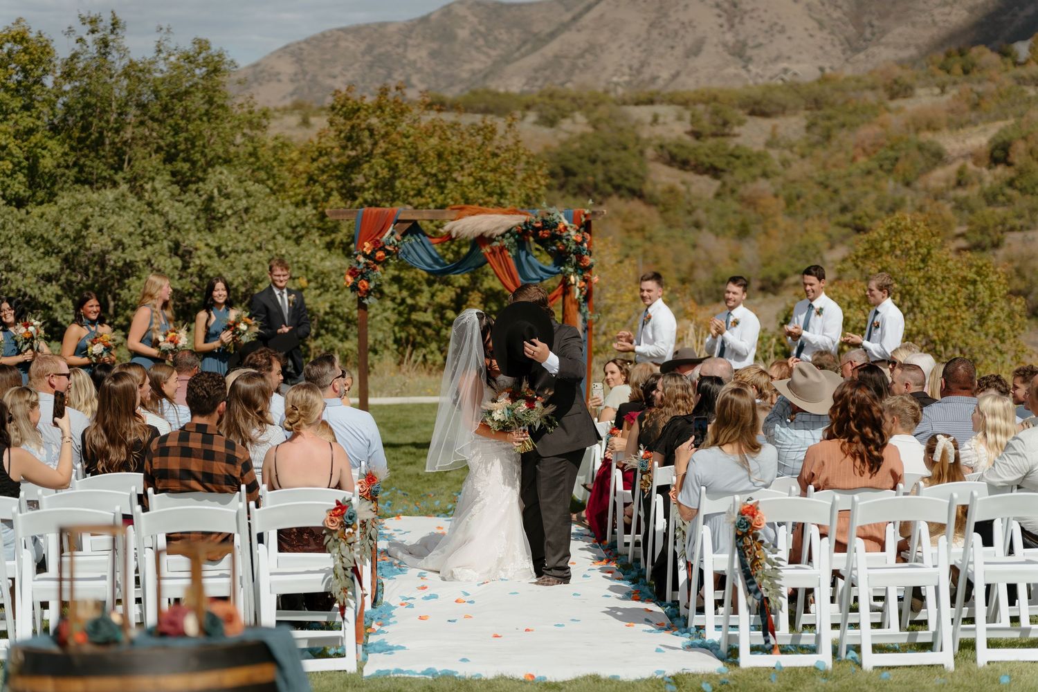 Outdoor wedding ceremony with bride and groom kissing at colorful floral arch, guests seated in white chairs, mountain backdrop.
