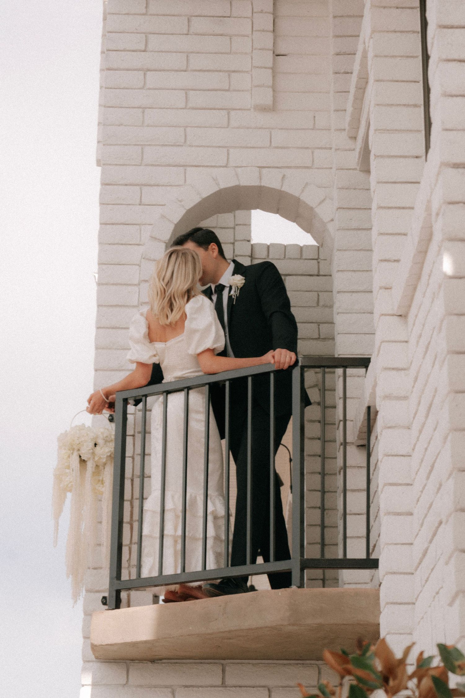 Wedding couple sharing a romantic kiss on a white brick building balcony with floral bouquet.