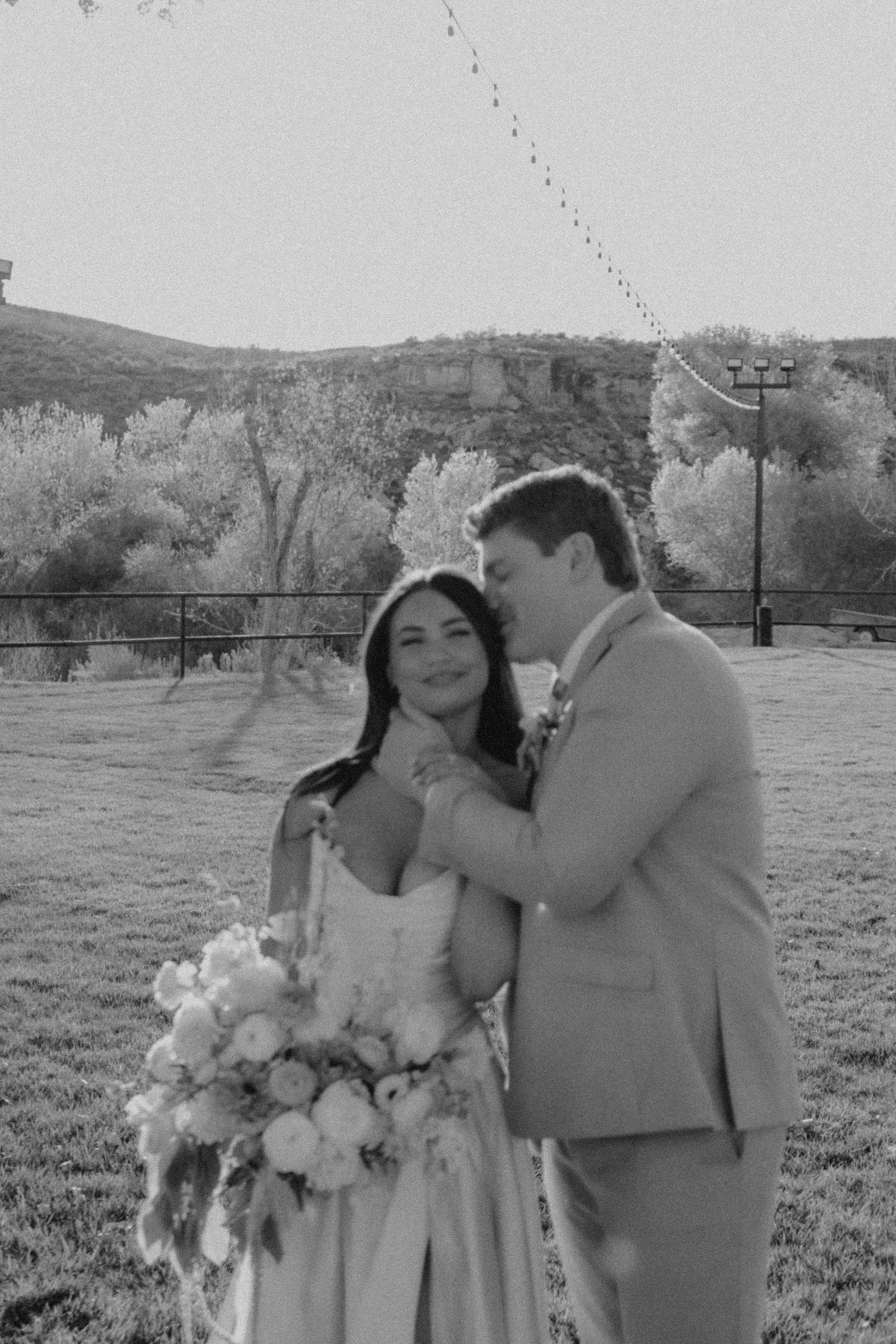 Black and white photo of a bride and groom embracing outdoors at a scenic wedding venue with trees and hills.
