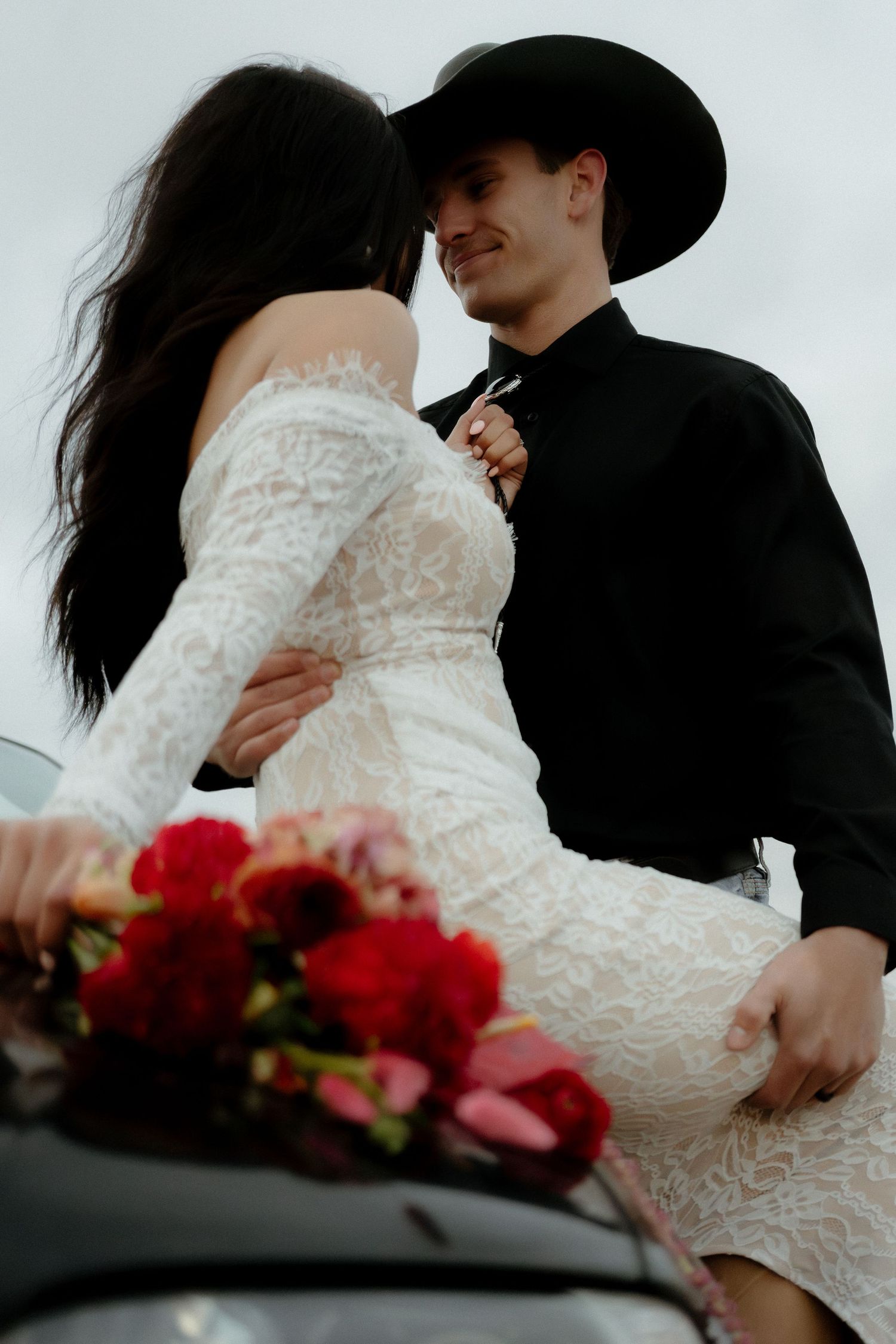 A bride in a lace dress and groom in black cowboy hat embrace romantically beside a car with red roses.