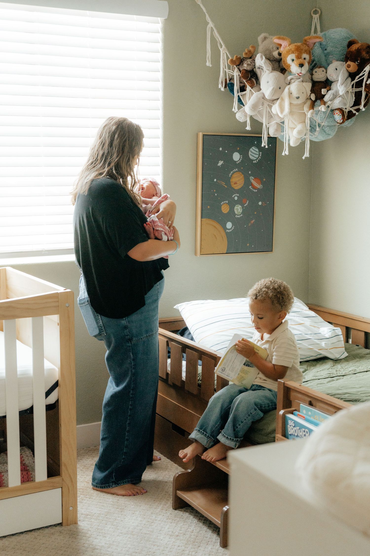 Mother holding newborn baby in nursery while toddler reads on wooden bed, with stuffed animals displayed above.