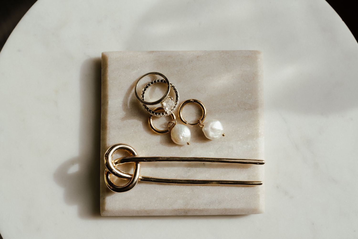 Gold rings and pearl earrings arranged on a marble tray with a gold hair pin on a white surface.