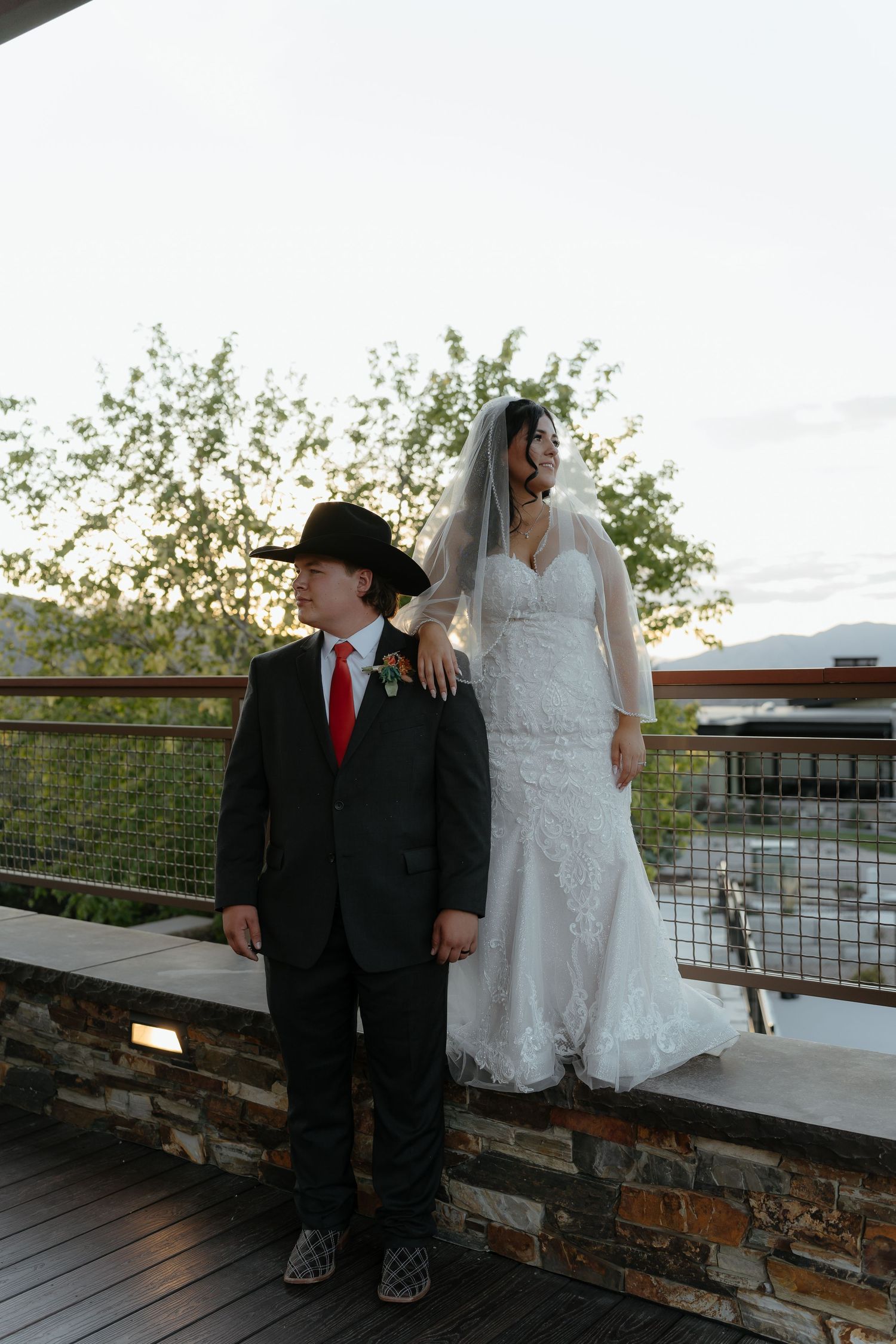 Bride in lace gown and veil stands on stone terrace with groom in black cowboy hat and suit at outdoor wedding.