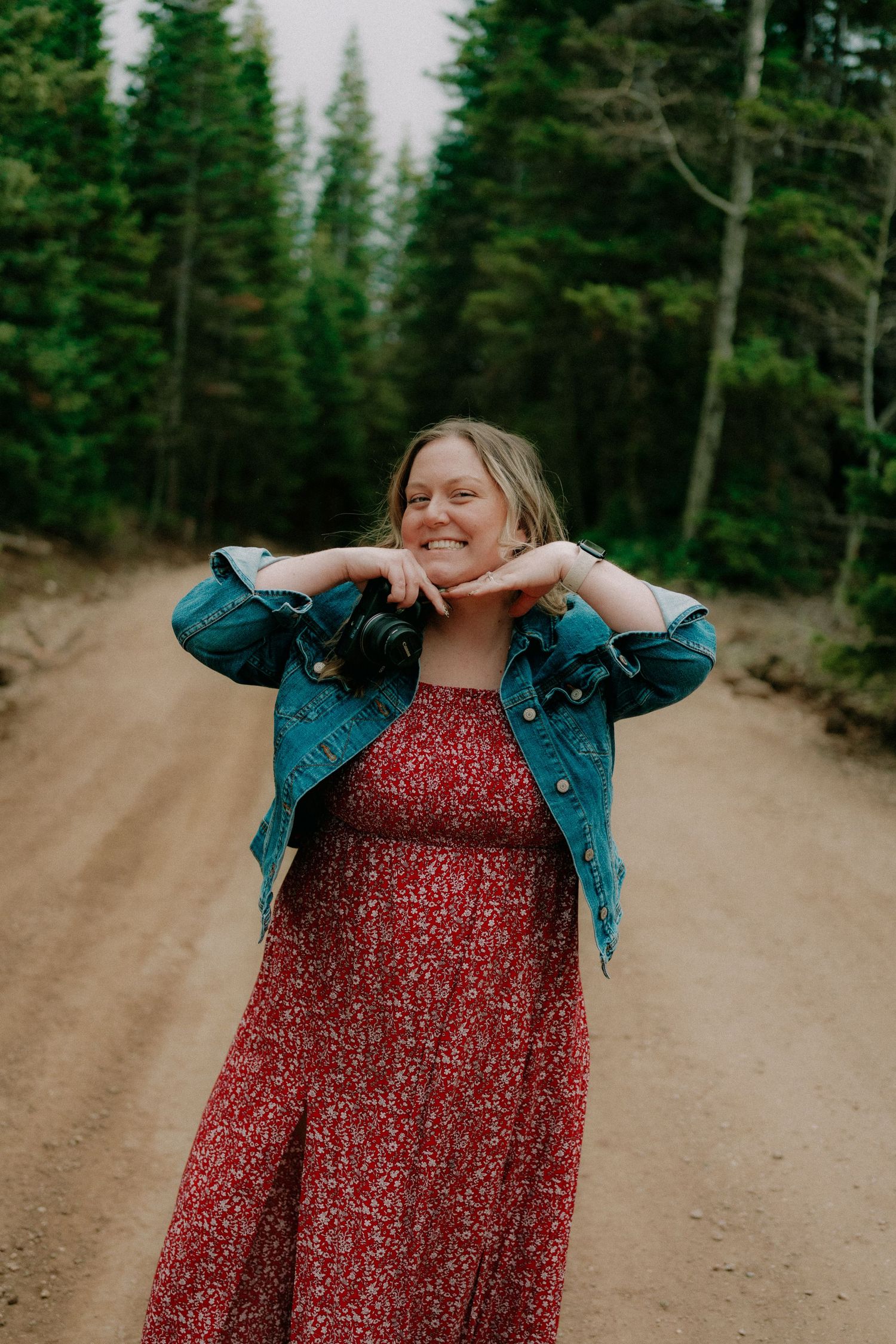 Woman in red floral dress and denim jacket smiling on a dirt forest road surrounded by tall evergreen trees.