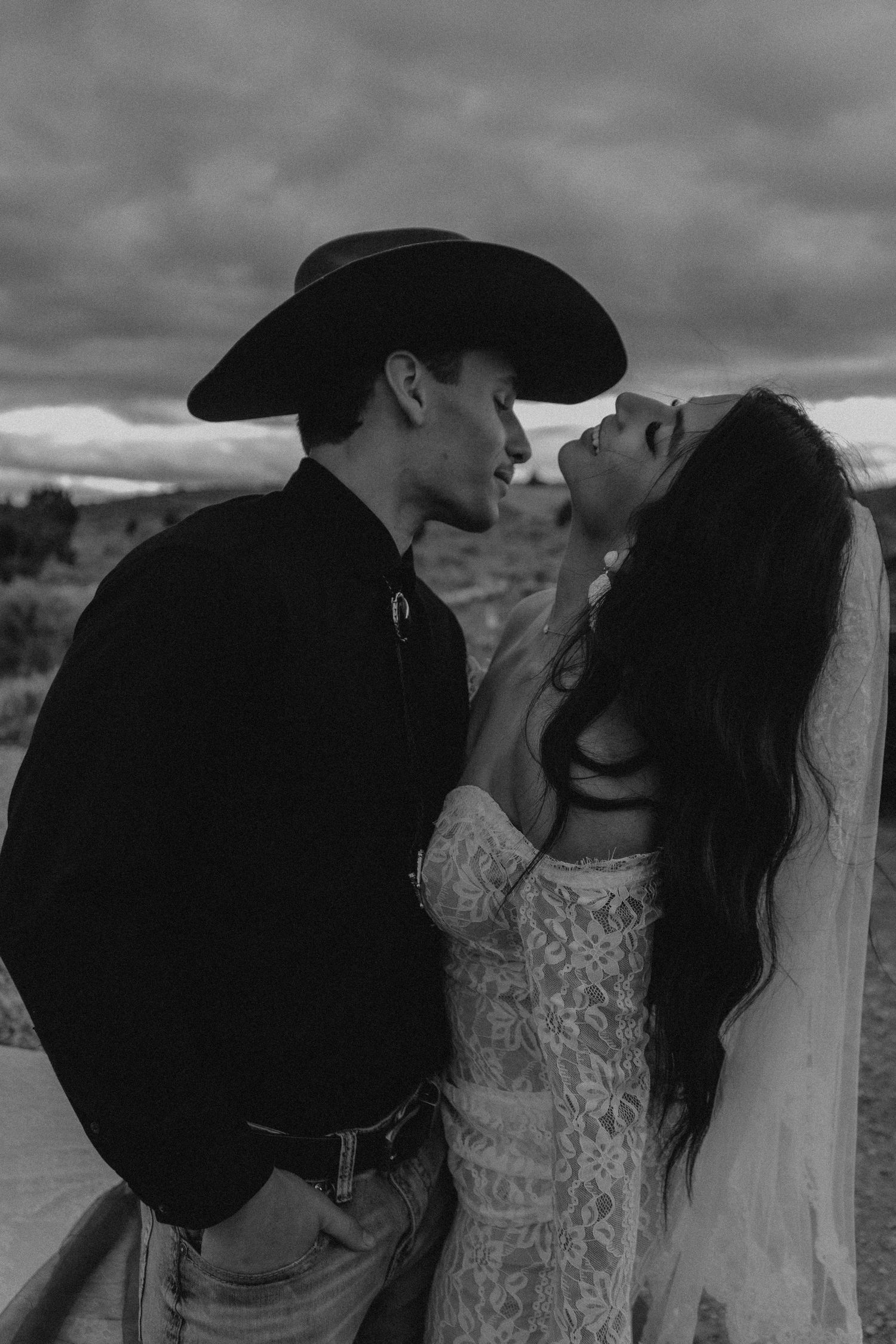 Black and white photo of a couple in western wedding attire sharing a romantic moment under dramatic cloudy skies.
