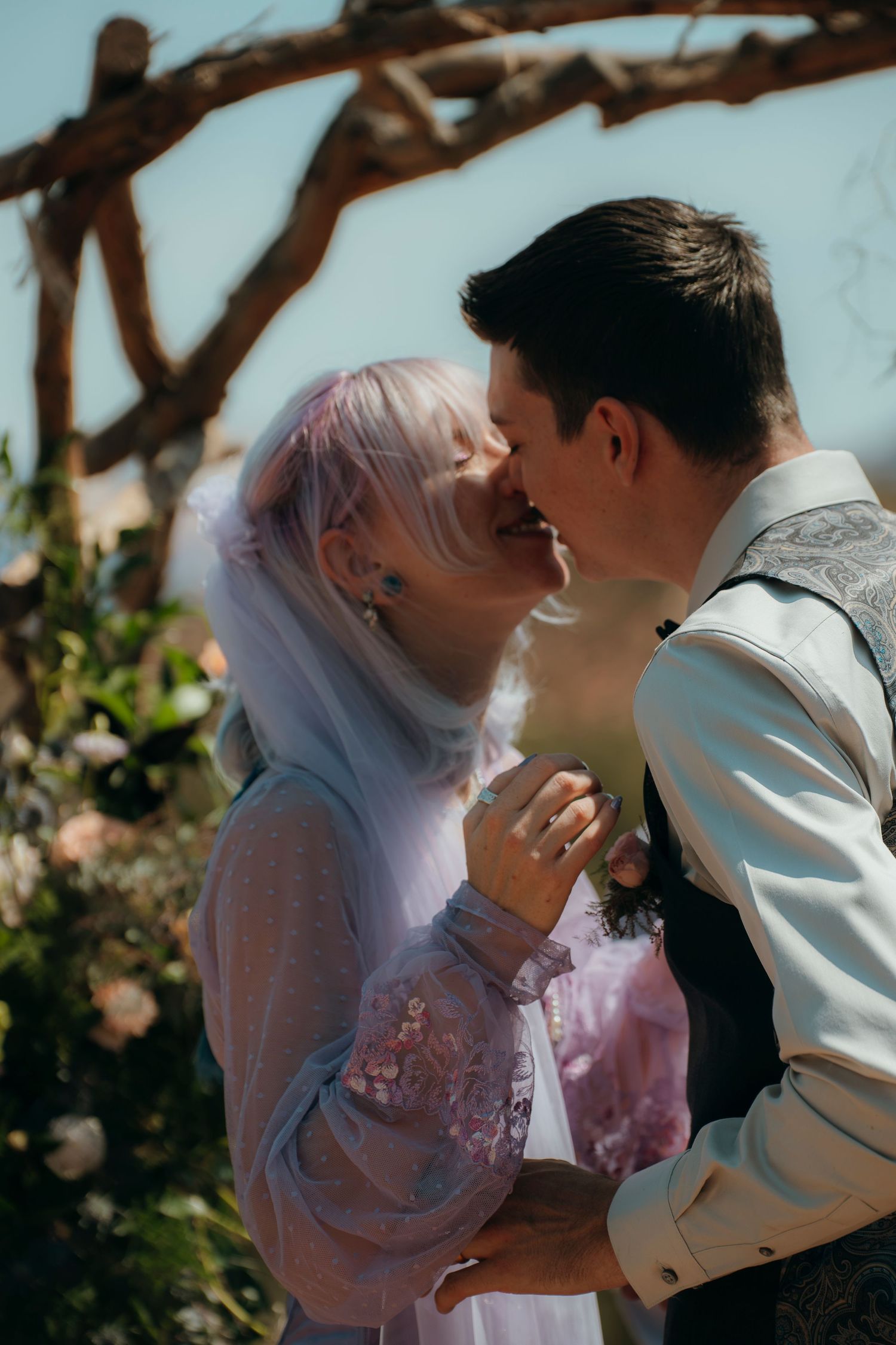 A bride and groom share a romantic kiss under a rustic wooden arch adorned with pink flowers at an outdoor wedding.