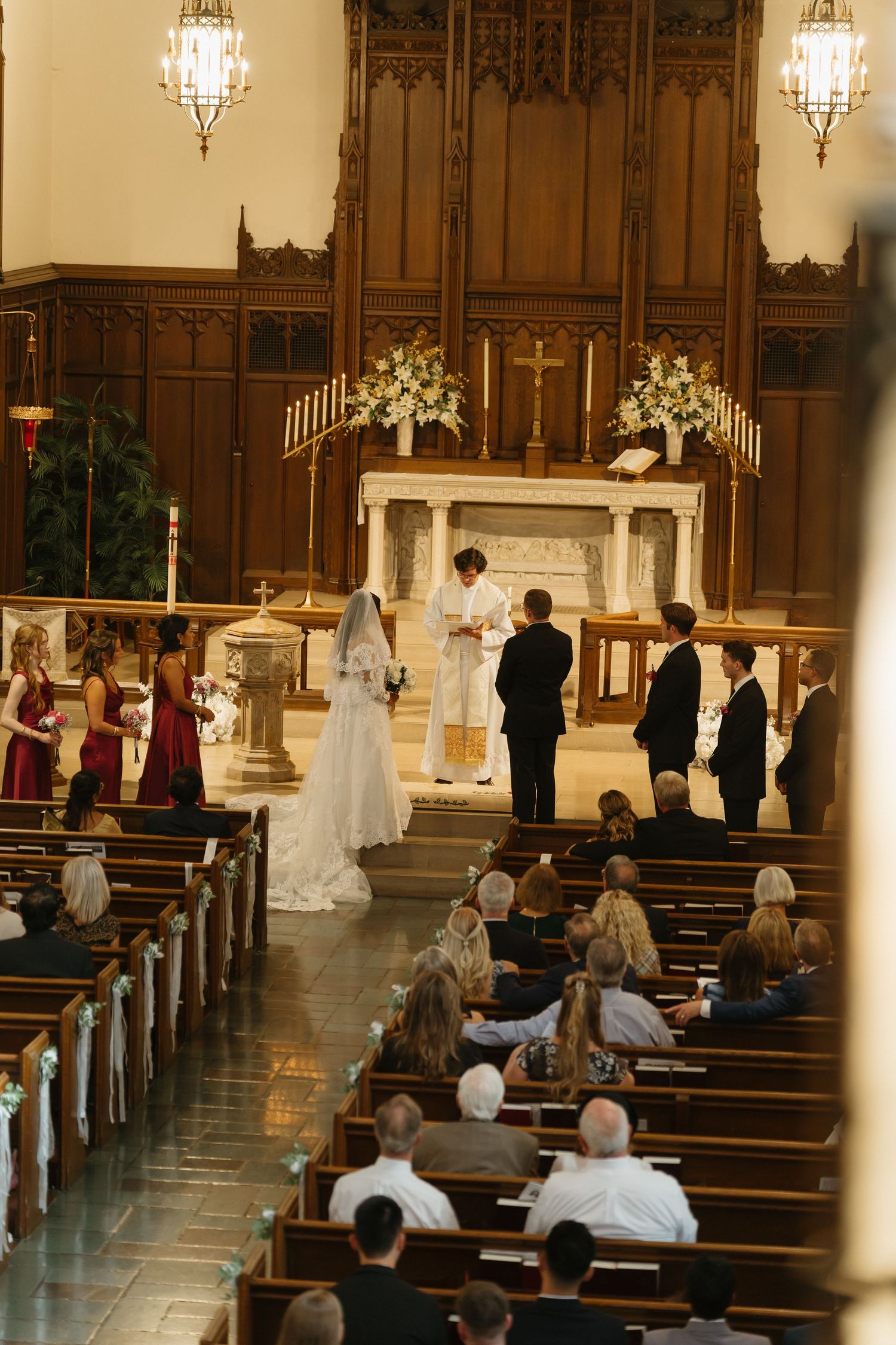 A bride and groom exchange vows at a church altar surrounded by groomsmen and bridesmaids with guests seated in wooden pews.