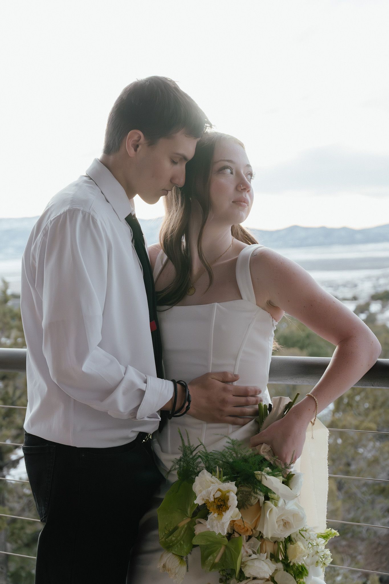 Groom embraces bride from behind as she holds white bouquet on outdoor balcony with scenic view.