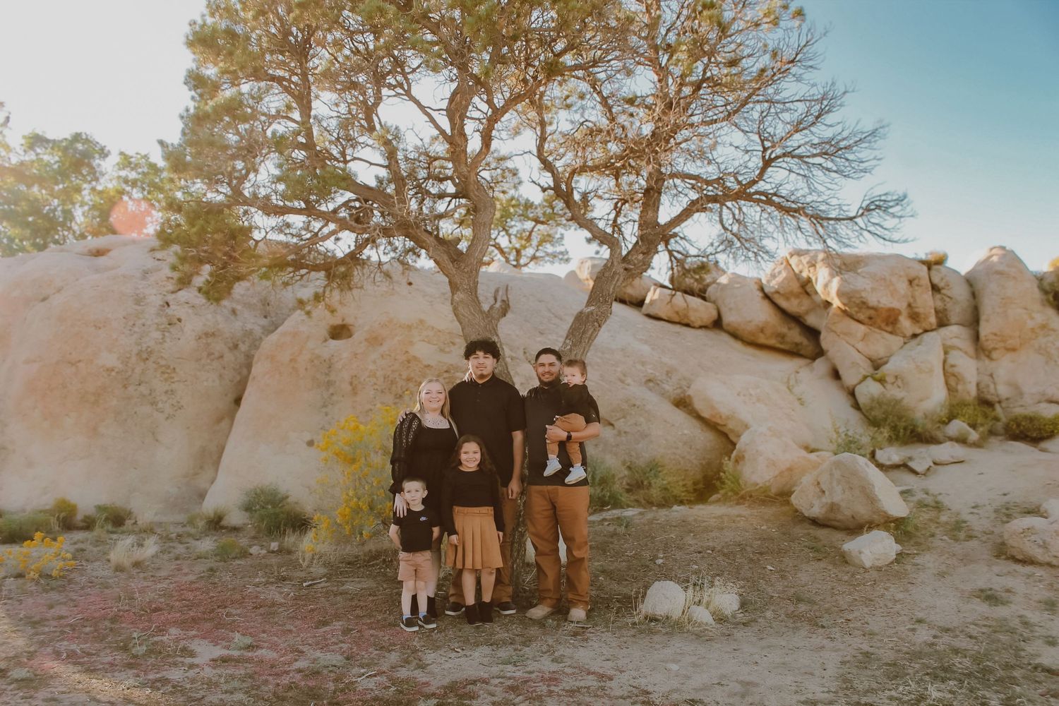 Family of six posing among large boulders and a tree in a desert landscape at golden hour.