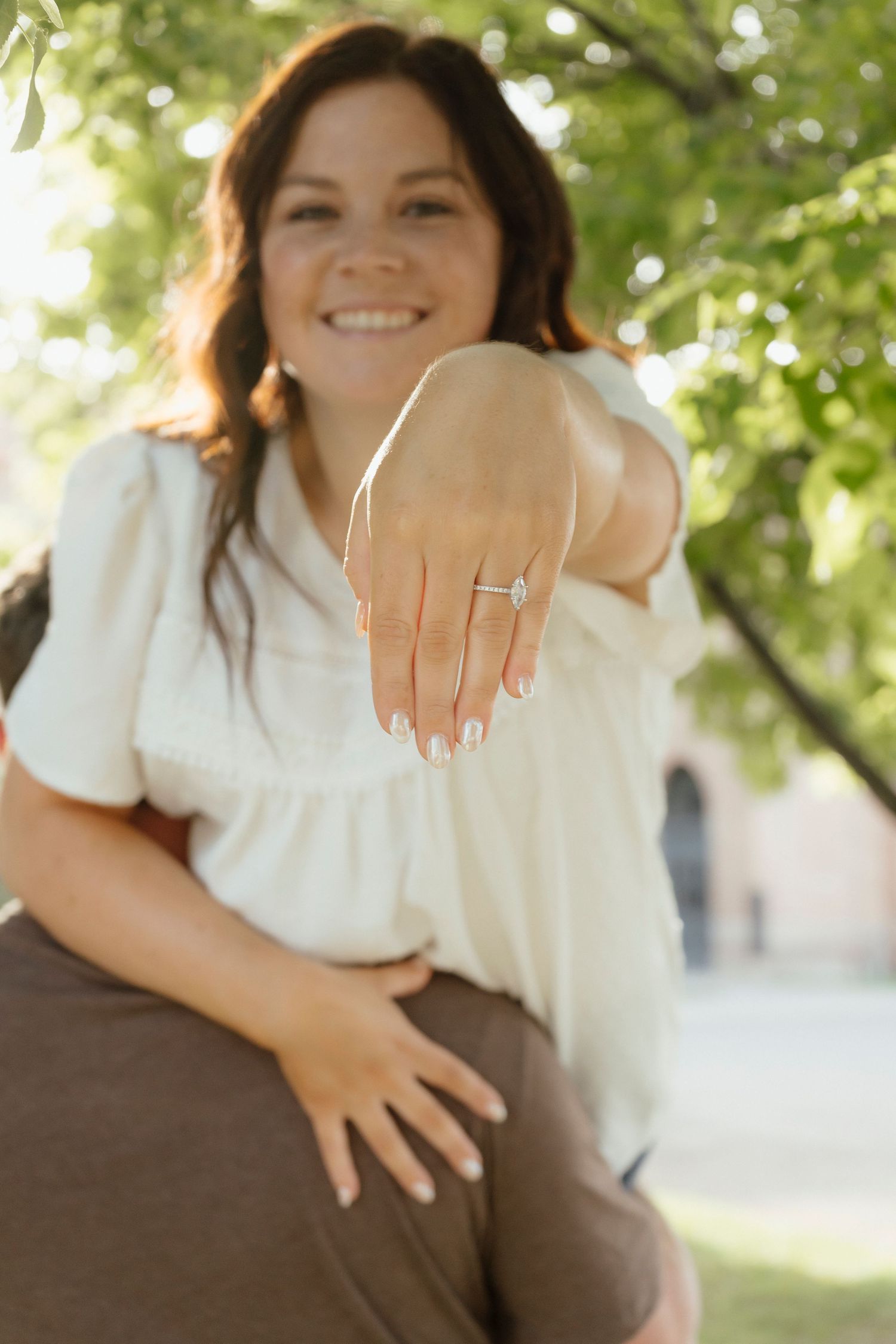 Smiling woman outdoors showing off her engagement ring, extending hand toward camera near green trees.