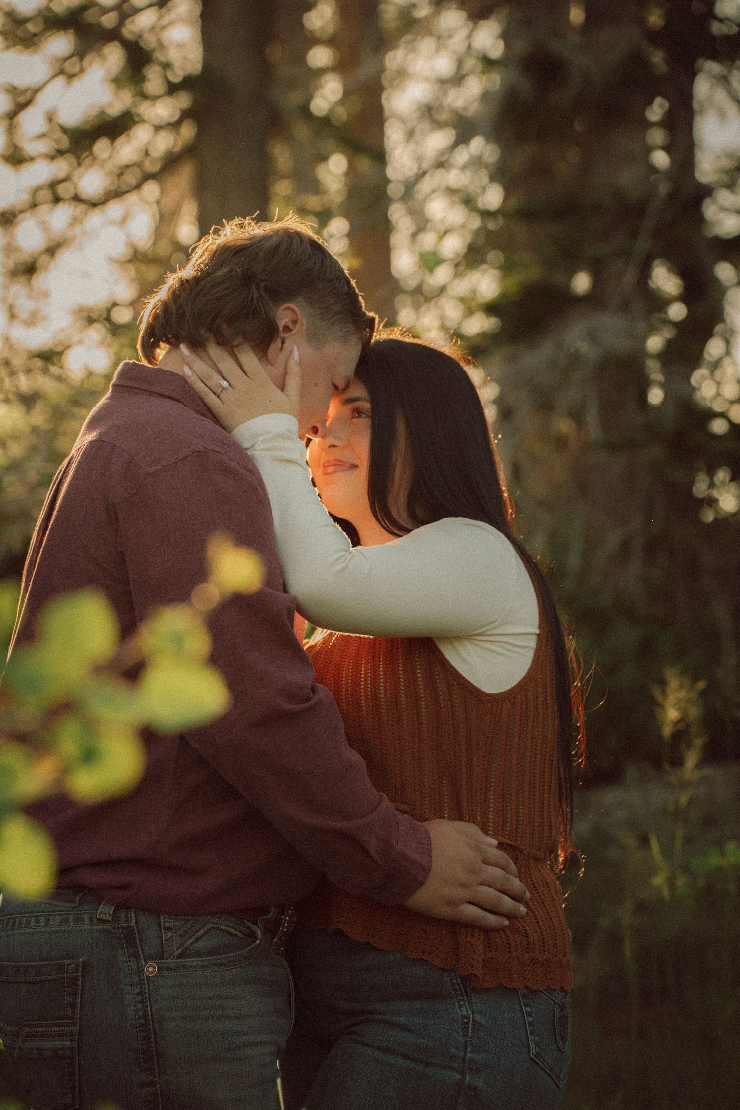 Couple embracing and kissing at golden hour in a forest, warm sunlight glowing behind them.