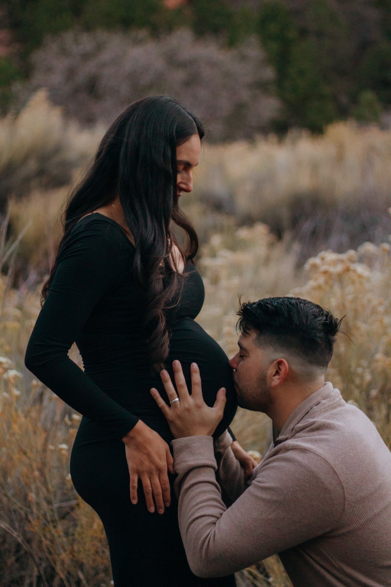 Man kneeling to kiss pregnant woman's baby bump in a scenic outdoor field with dry brush.
