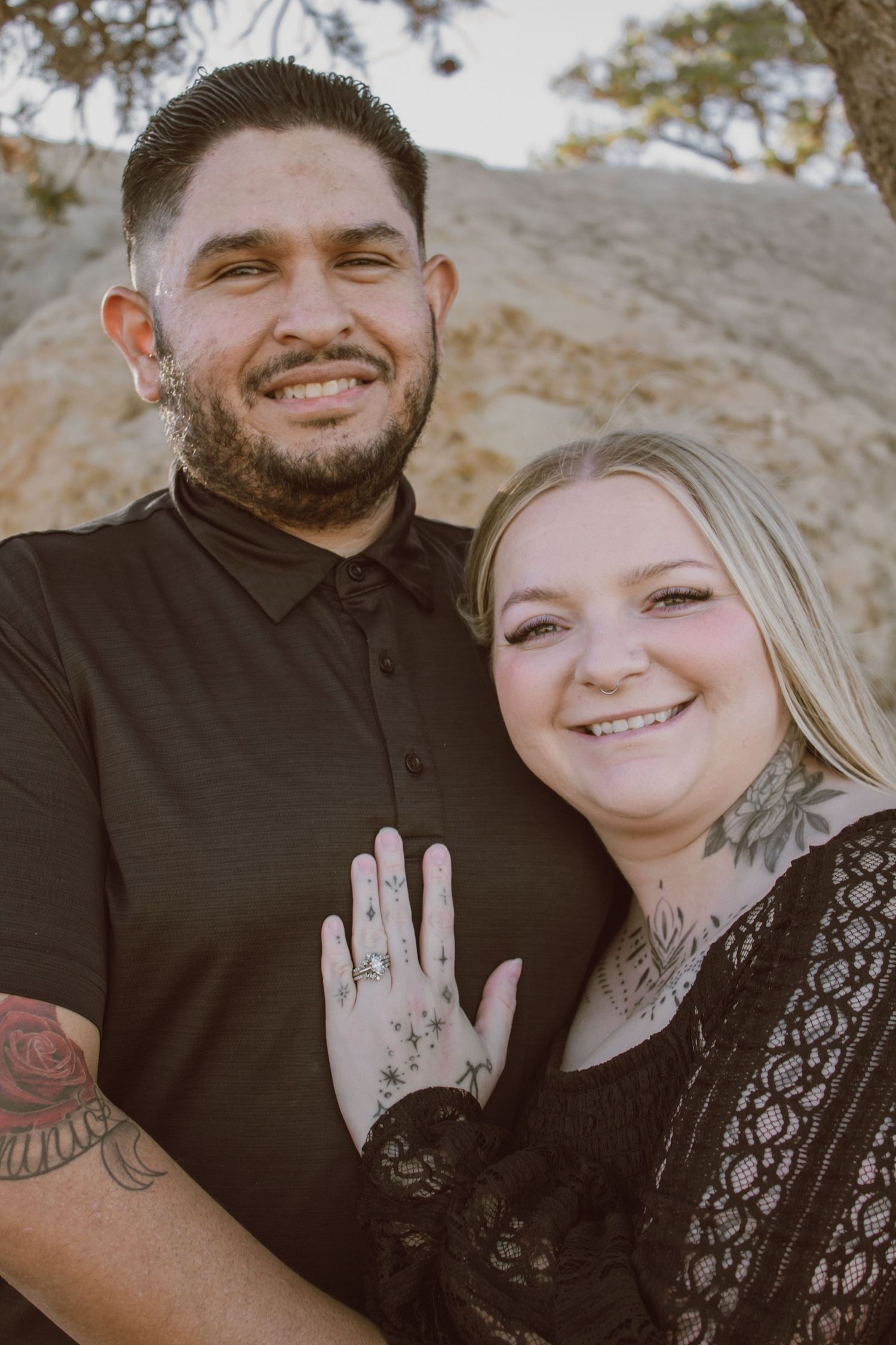 Smiling tattooed couple showing engagement ring outdoors with rocky background in warm golden light.