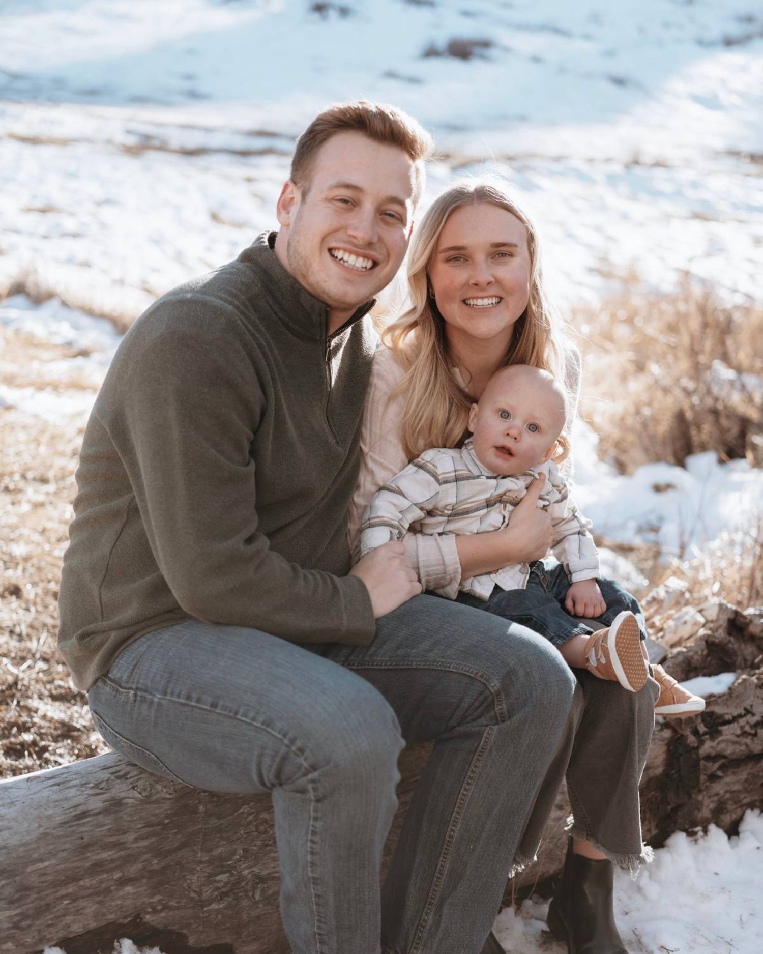 A smiling young couple sits on a snowy hillside log, holding their baby in a cozy winter family portrait.