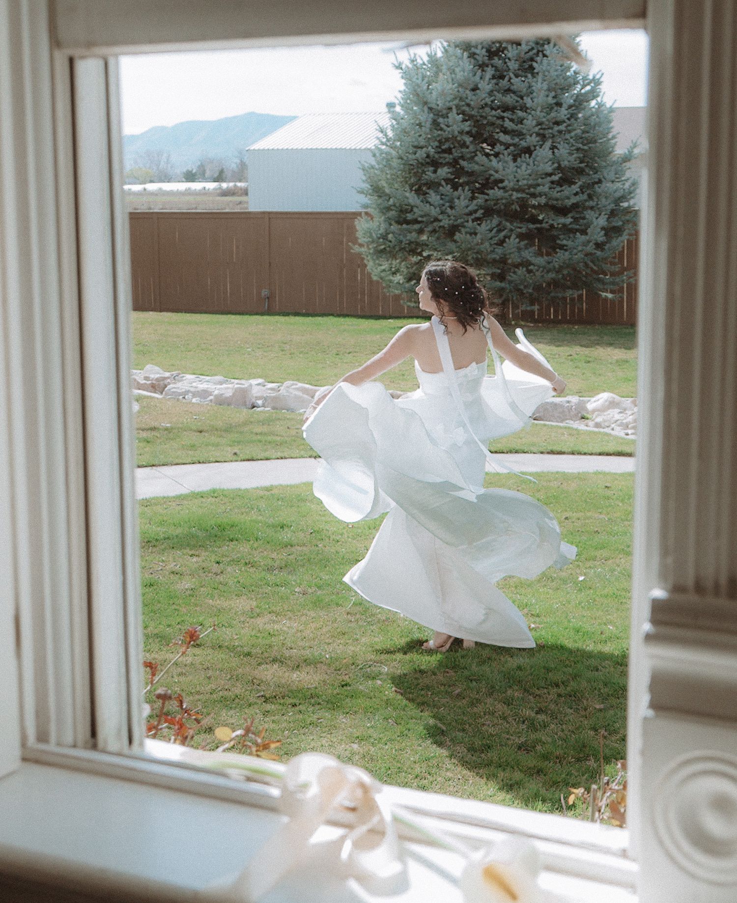 Bride in flowing white wedding gown twirling in a garden, viewed through a window frame.