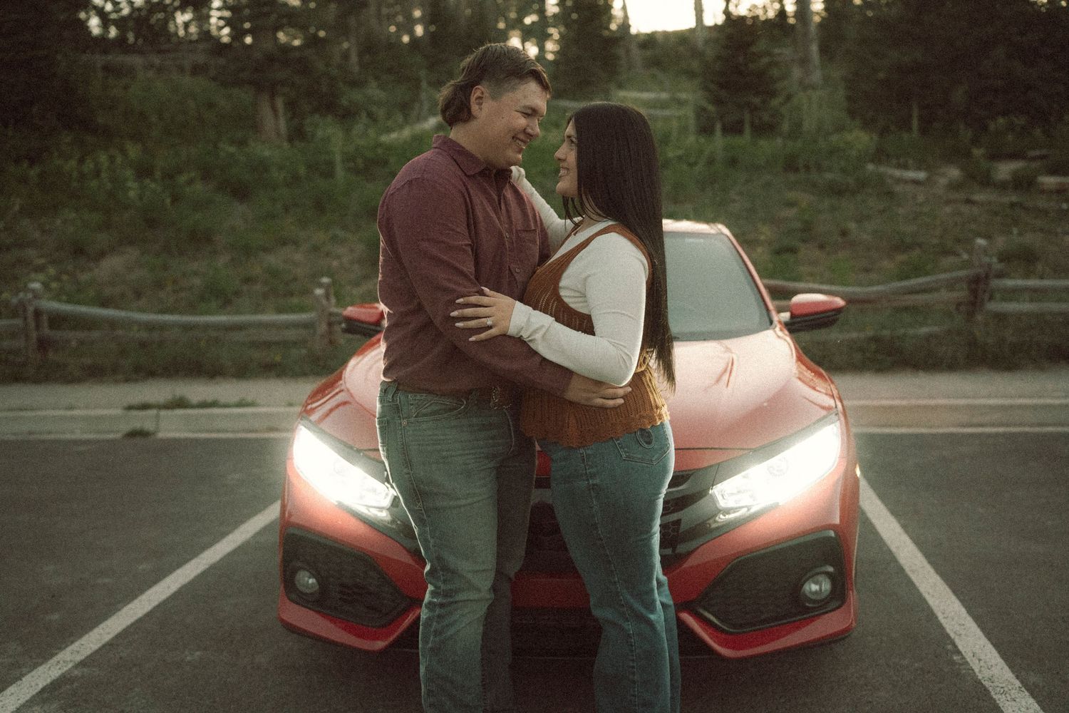 A couple embraces romantically in front of a red Honda Civic at dusk in a parking lot.